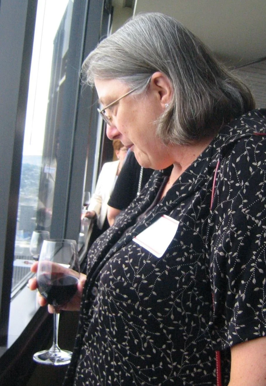 A woman with gray hair and glasses holding a glass of red wine, standing near a window during a professional event or conference.