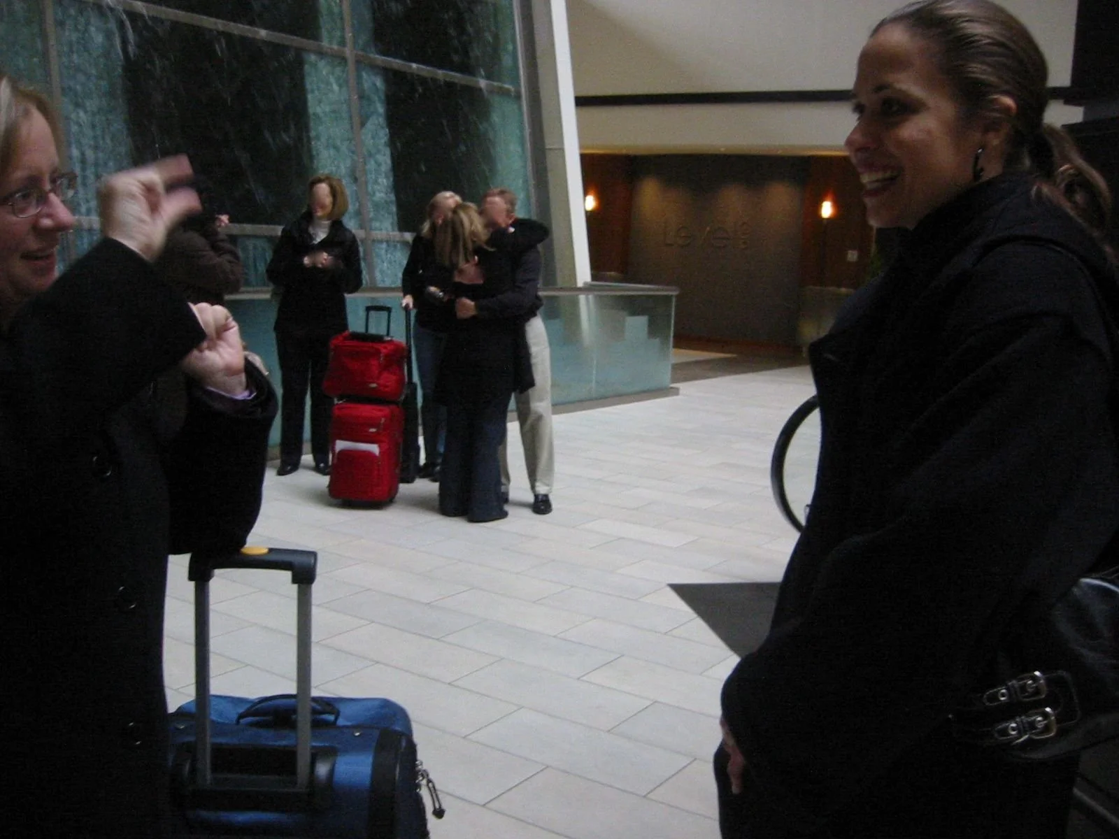 A group of people at an airport terminal, some engaged in conversation. Two women in the foreground are chatting, one with a luggage cart. In the background, a couple is hugging near a red suitcase, while others stand and wait.