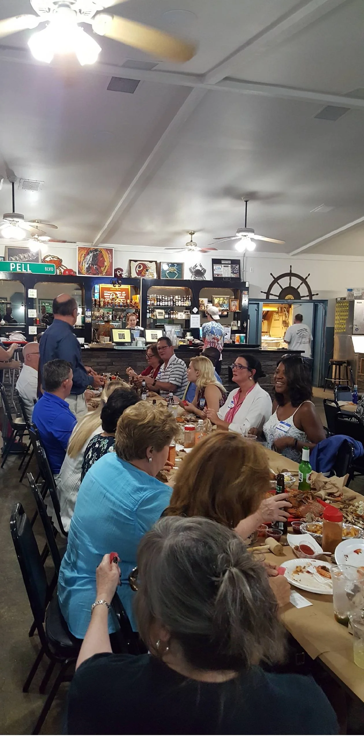 People sitting at a long table in a restaurant enjoying a meal with a crowd in the background, and a kitchen area visible behind the counter with staff preparing food. The room has white walls, ceiling fans, and decorative pictures and a ship wheel o