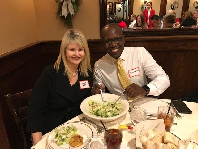 A man and woman sitting at a restaurant table, smiling at the camera, with a large bowl of salad and drinks on the table. The man is wearing glasses and a white shirt with a yellow tie; the woman has blonde hair and is wearing a black outfit. There i