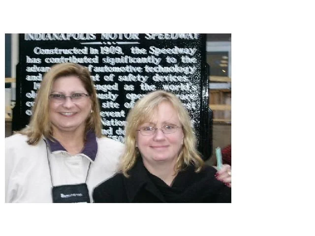 Two women smiling, one wearing glasses and a black top, the other with glasses and a white jacket, standing in front of a sign about Indianapolis Motor Speedway
