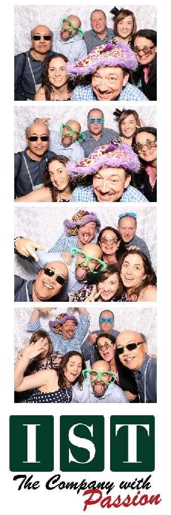 Group of people in a photo booth with props, wearing sunglasses, colorful hats, and funny glasses, having fun and making silly faces. The bottom features a logo with the letters IST and the phrase "The Company with Passion."