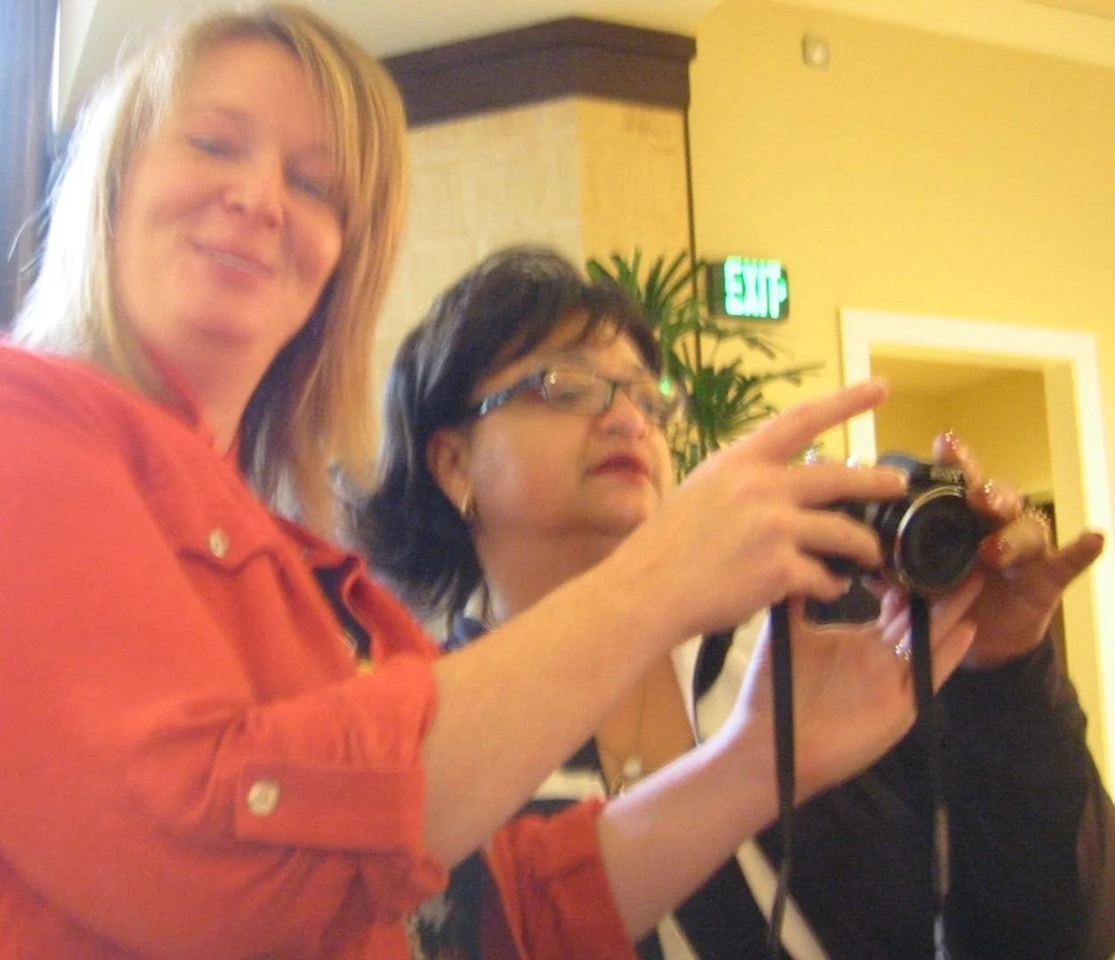 Two women in a room, one with blonde hair wearing an orange shirt, smiling at the camera, and the other with dark hair and glasses, focusing on a camera she is holding and pointing it at something.