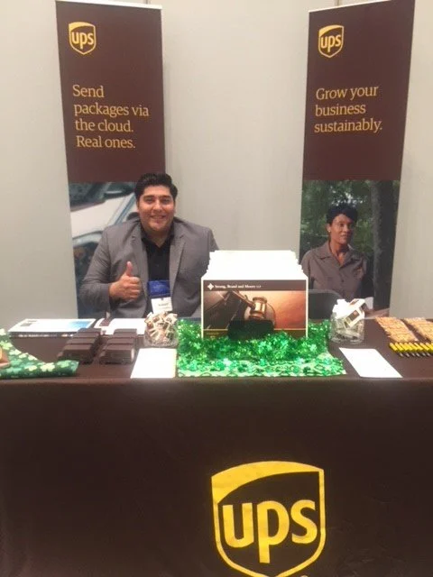 A person sitting at an indoor UPS promotional booth, smiling and giving a thumbs-up, with promotional banners behind. The booth has UPS branding and some giveaway items on the table.