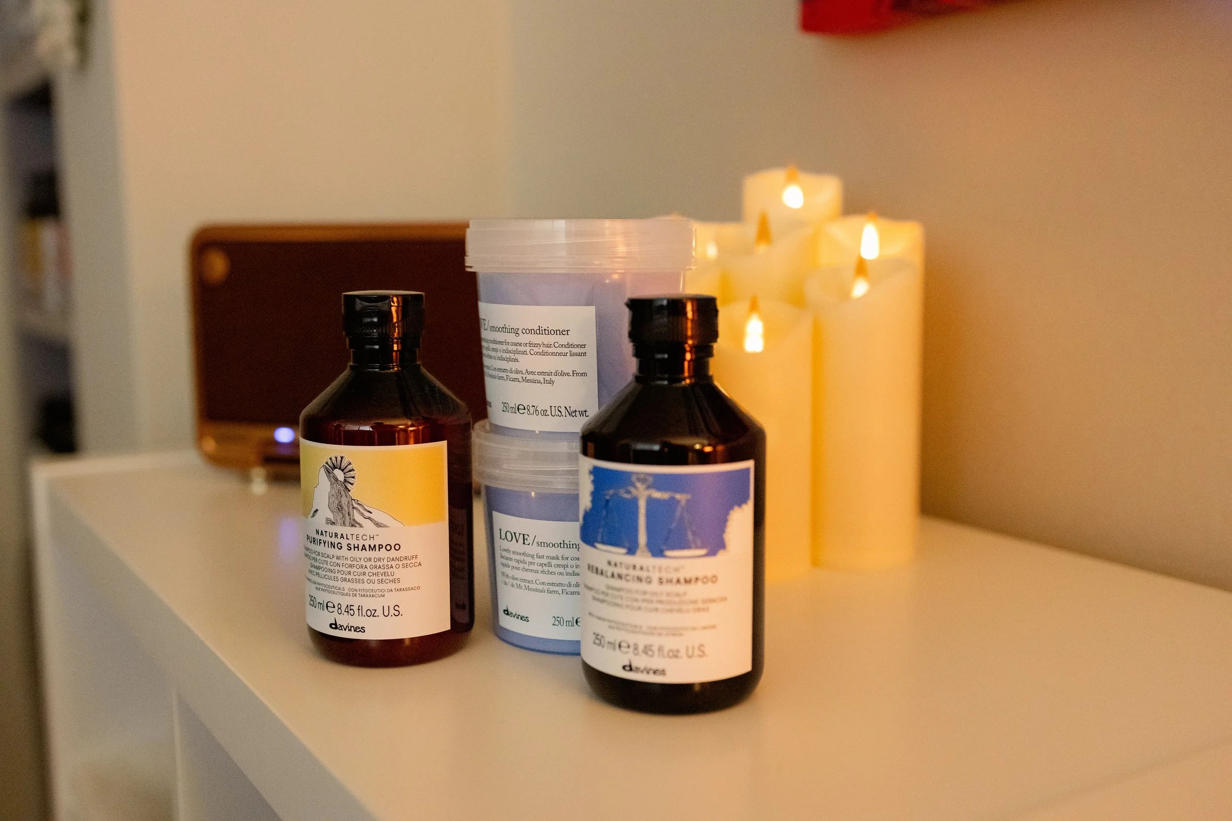Varios frascos de productos para el cabello, incluyendo shampoo y acondicionador en una repisa blanca, con velas encendidas en el fondo.