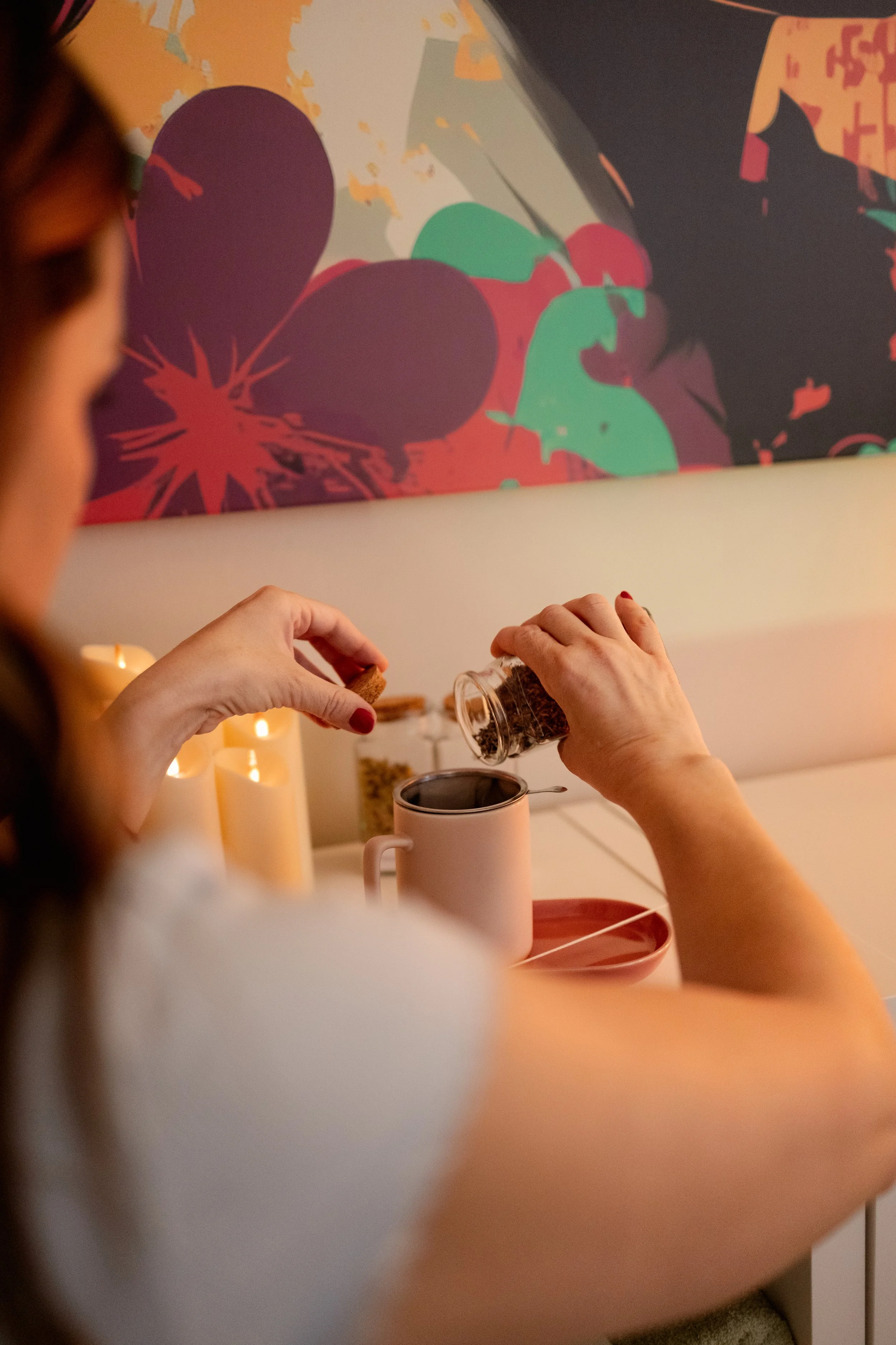 Manos vertiendo café de un frasco en una taza blanca en un ambiente cálido con velas y una decoración colorida en la pared.