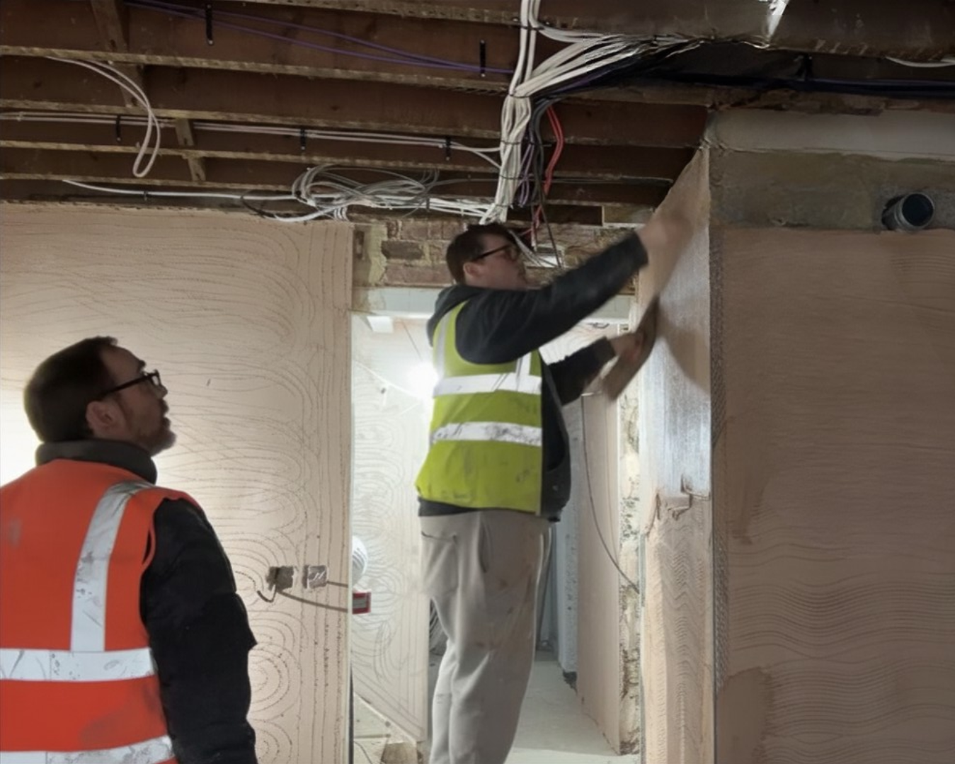 Two construction workers inspecting an interior space with exposed ceiling pipes and wires, one wiping a wooden surface and the other observing.