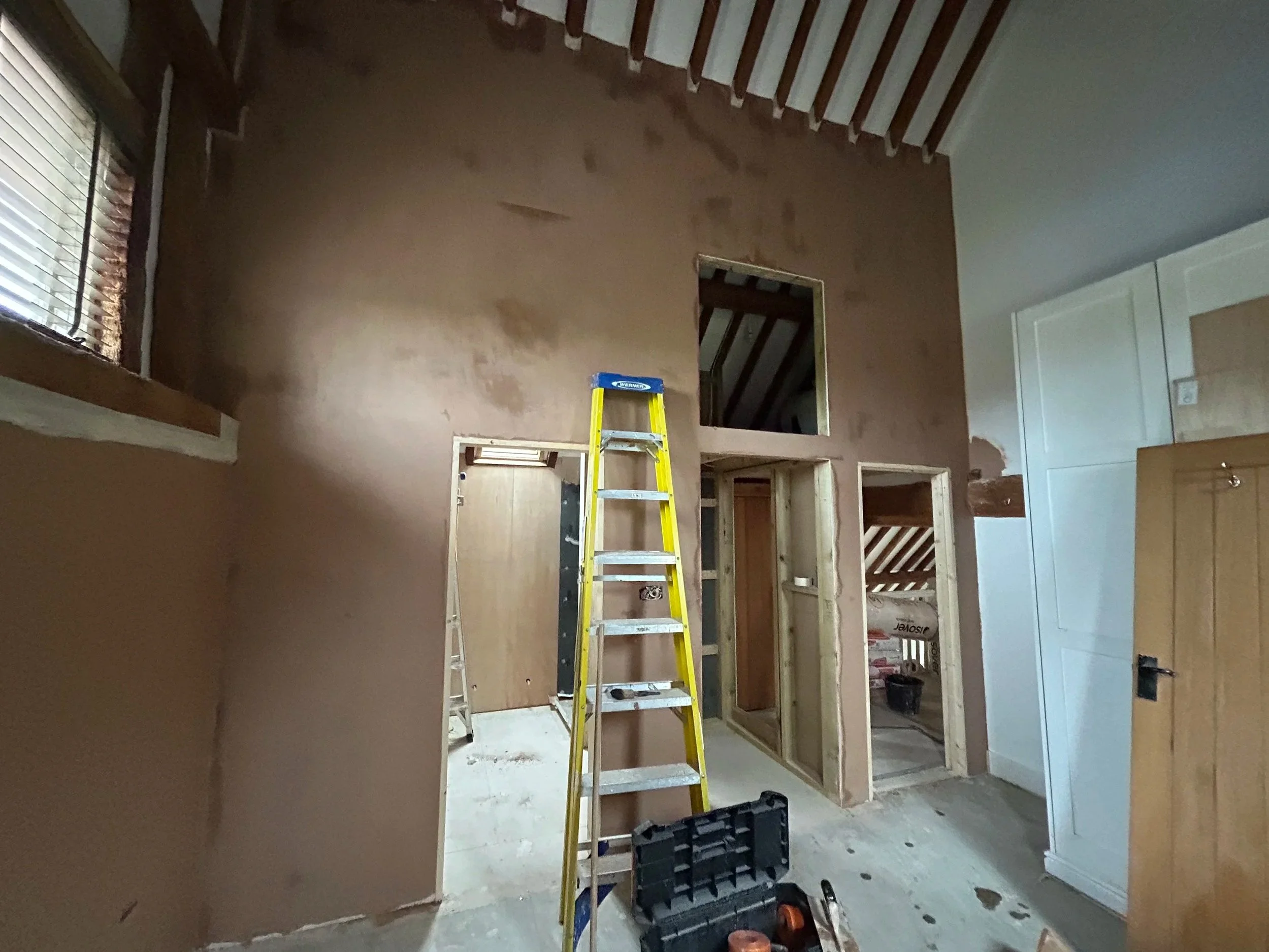 Interior of a room under construction or renovation with a ladder and partially finished walls, including a window with blinds and exposed doorway framing.