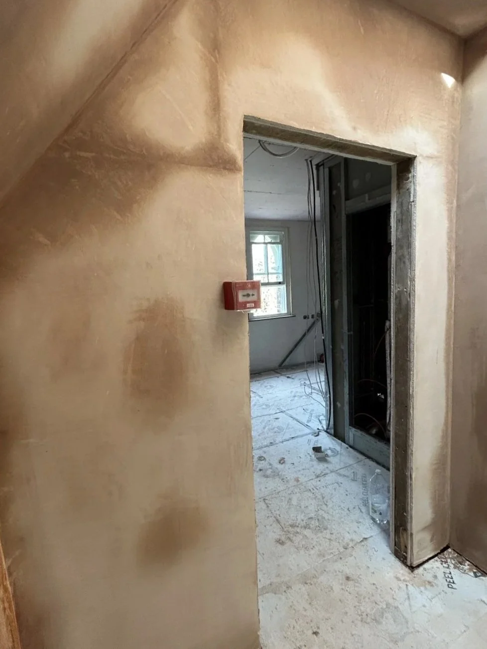 Interior view of a building under renovation, showing a doorway without a door, exposed wall studs, electrical wires, a window in the background, and construction debris on the floor.
