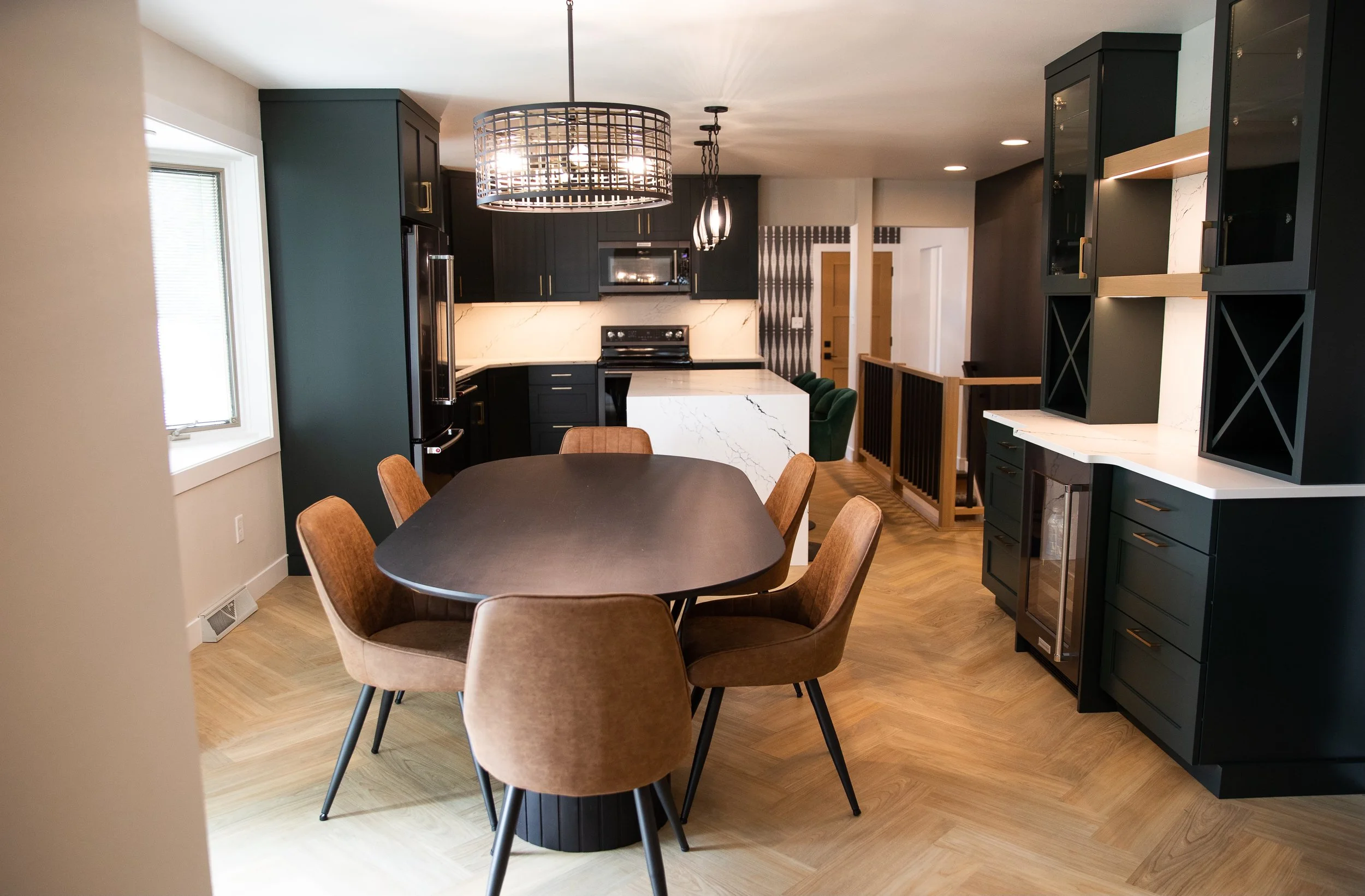 Modern kitchen with dark cabinetry, white marble countertops, and wooden flooring, featuring a round dining table with six brown chairs and decorative light fixtures.