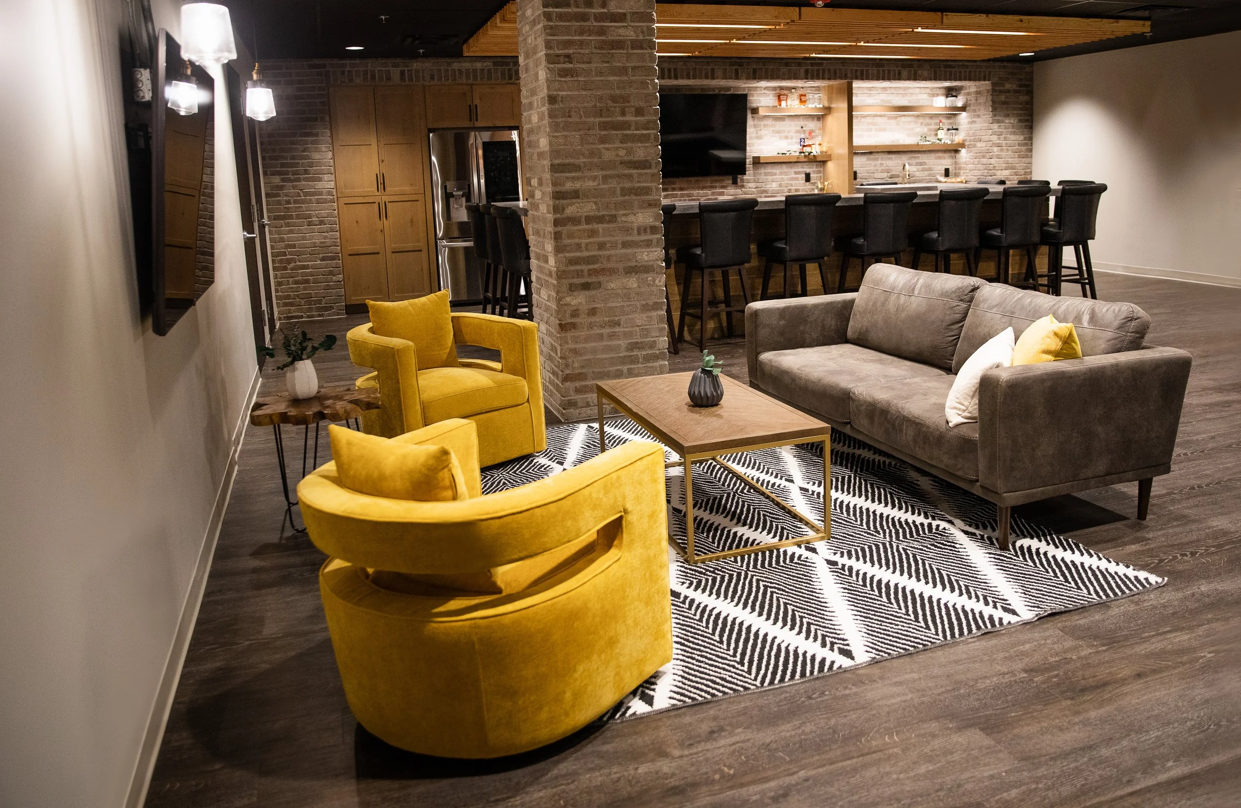 A modern living room with yellow armchairs, a gray sofa, a patterned black and white rug, and a wooden coffee table. In the background, there is a kitchen bar with black chairs, a brick wall, and wooden shelves.