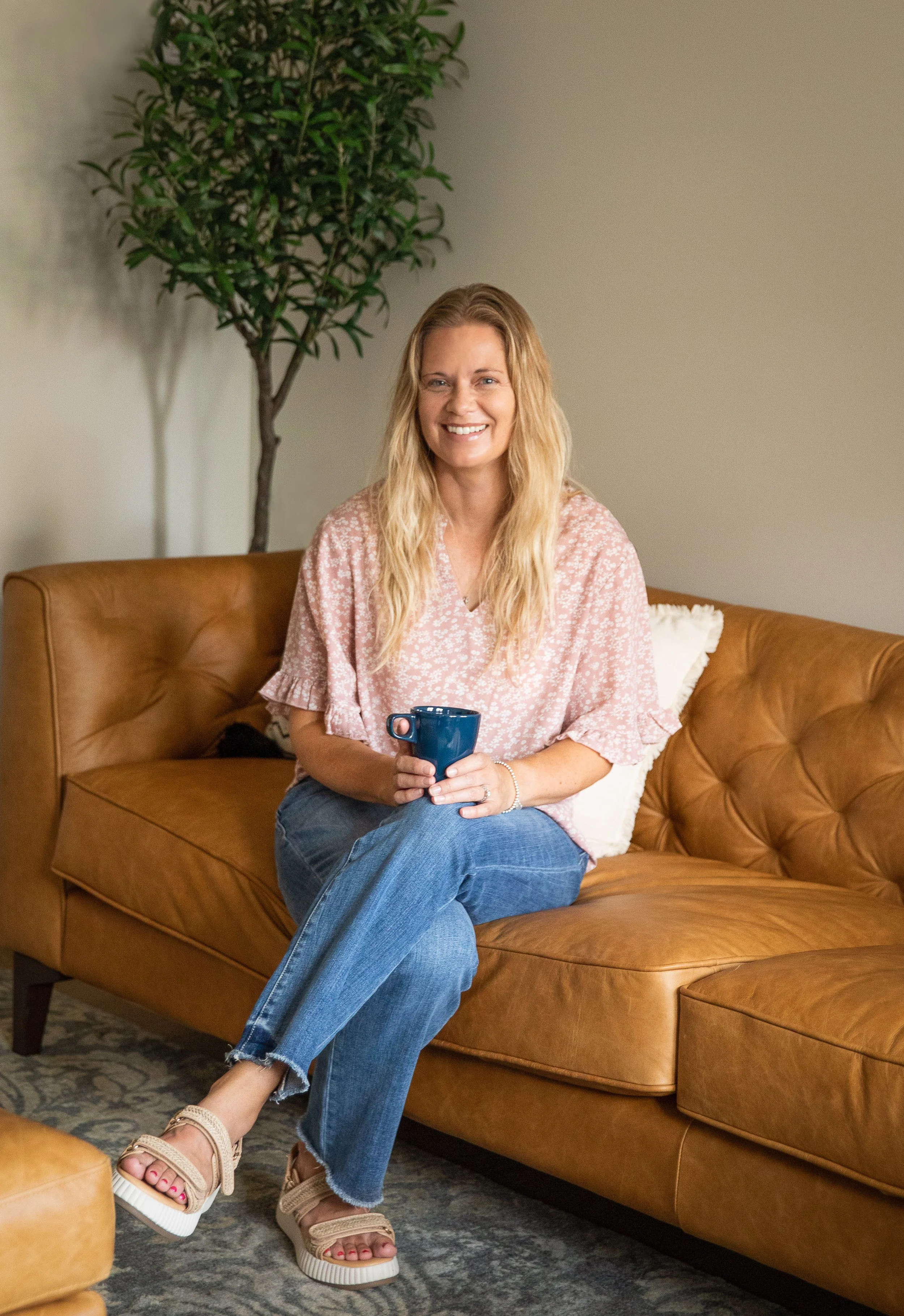 A woman with long blonde hair, wearing a pink patterned blouse and blue jeans, sitting on a tan leather sofa holding a dark blue mug, smiling at the camera. There is a tall green potted plant in the background and a decorative pillow on the sofa.