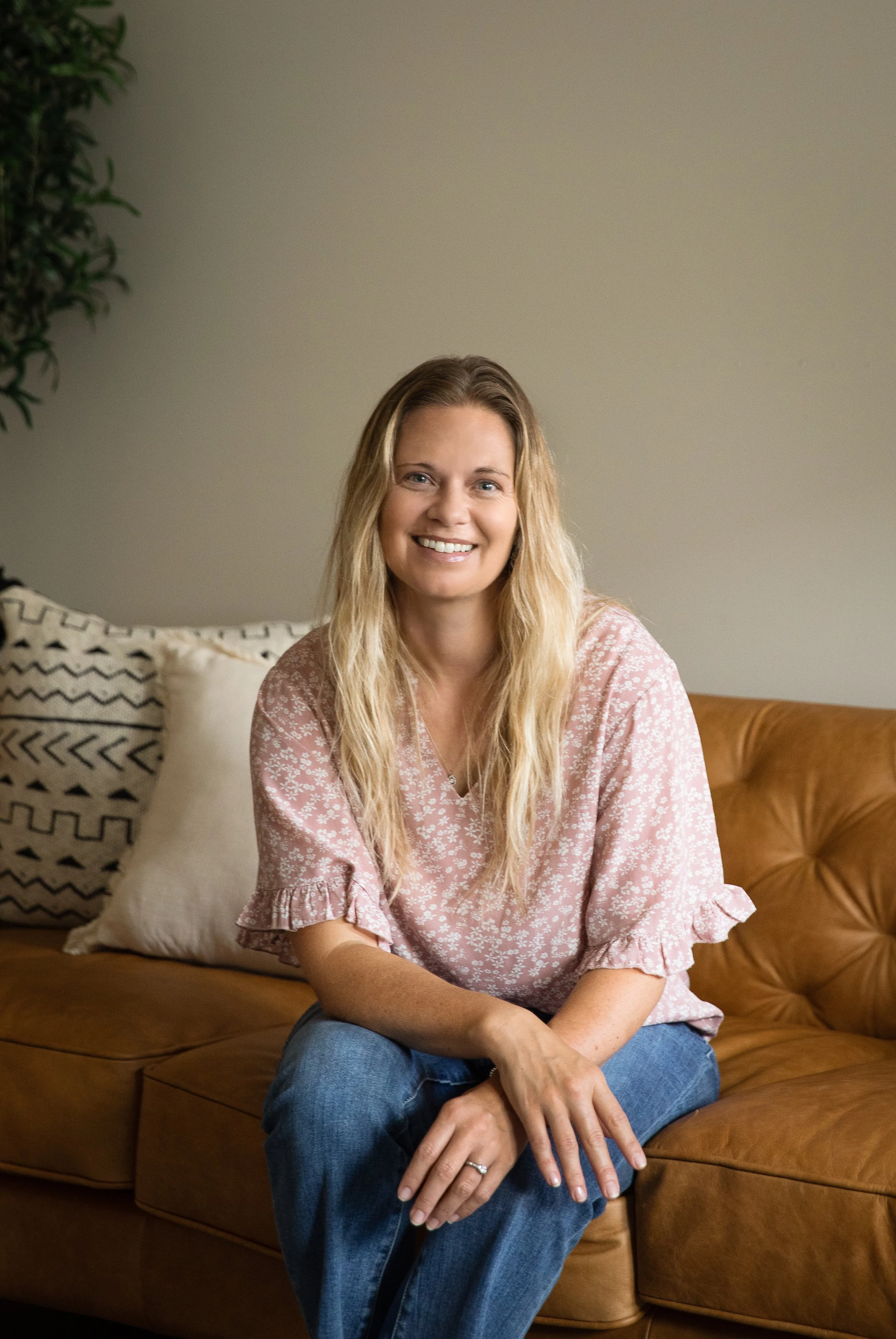 A woman with long blonde hair and a pink floral blouse smiling and sitting on a brown leather couch in a cozy living room with decorative pillows and a green plant in the background.