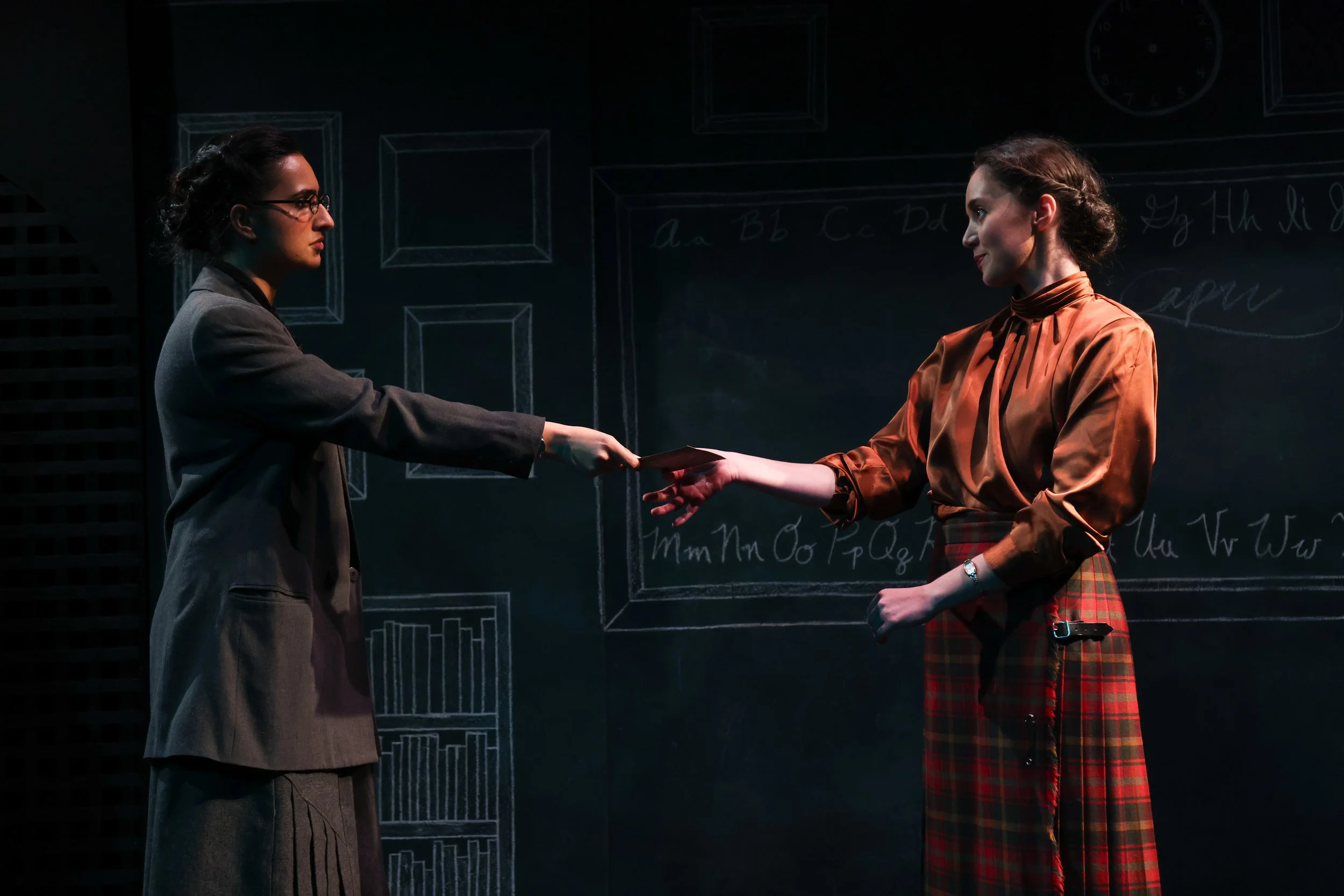 Two women are on stage, one on the left in a gray suit with glasses, facing the other on the right, who is wearing a brown satin blouse and a red plaid skirt. The woman on the right is handing an object to the woman on the left. The background featur