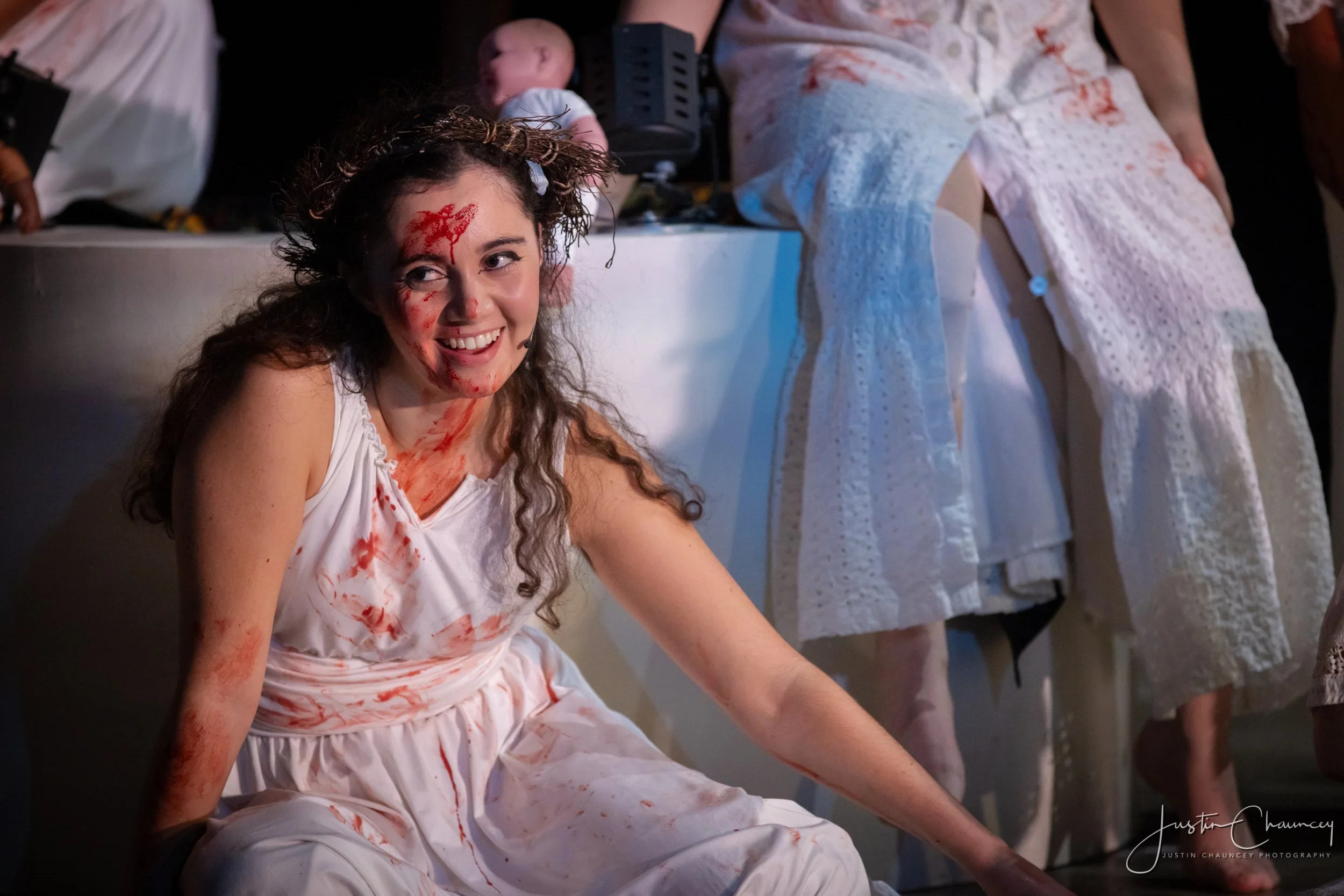 A young woman with curly hair, wearing a white dress, smiling and covered in fake blood, sitting on the floor in a dimly lit room with dresses hanging behind her.