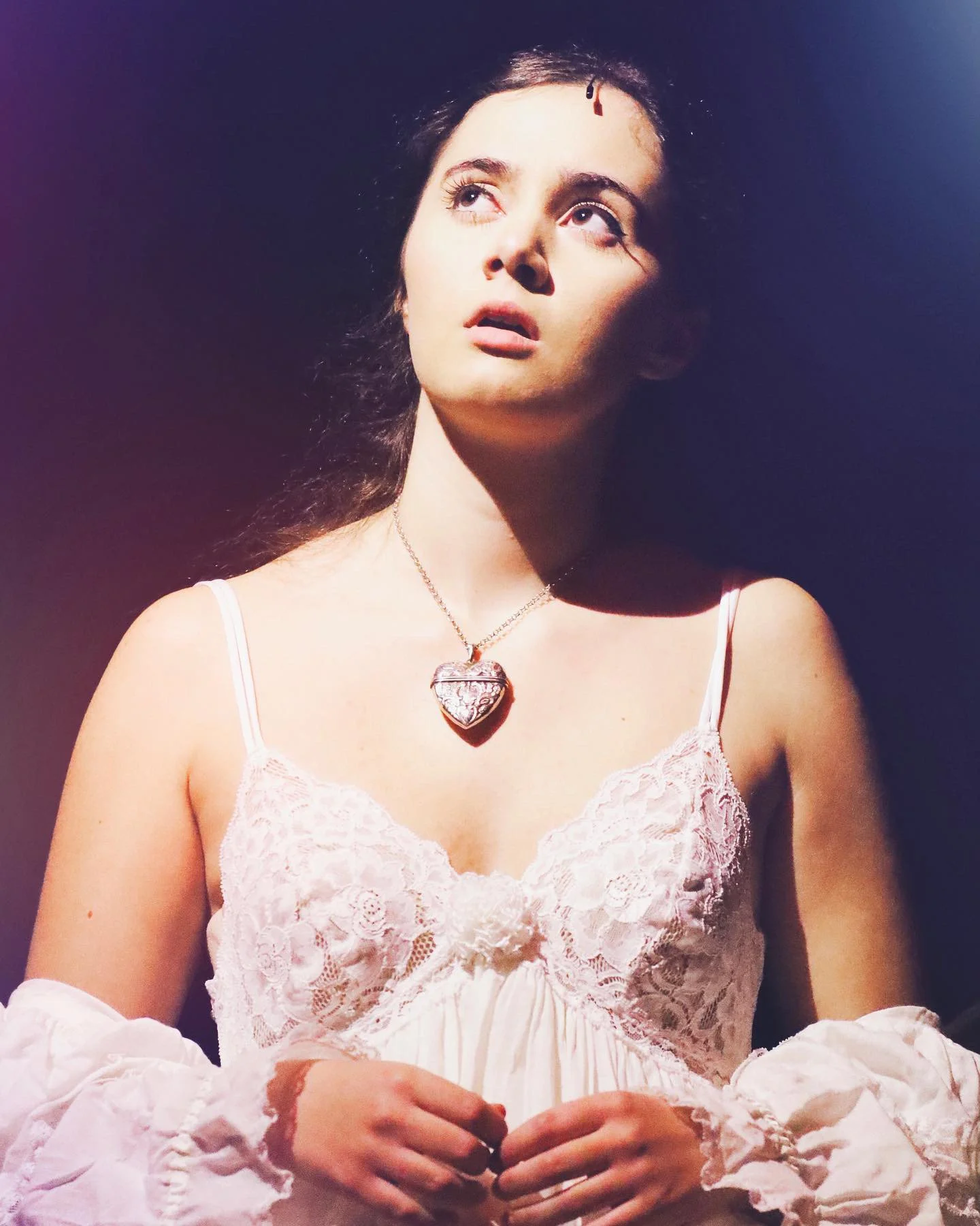 A young woman with long dark hair and light skin wearing a pink lace off-shoulder dress and a heart-shaped necklace, looking upwards with a thoughtful expression, against a dark background.