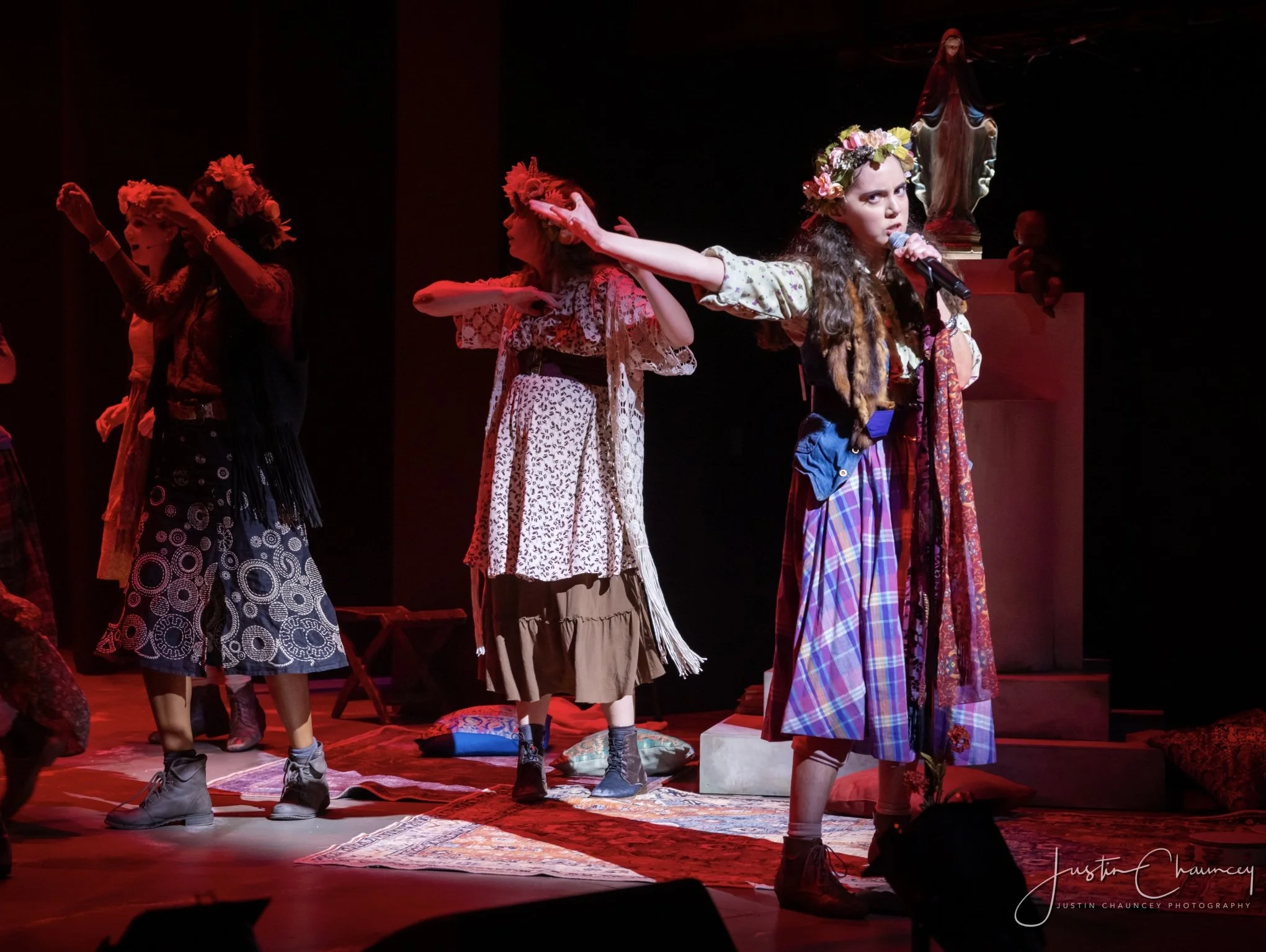 Children dressed in traditional and colorful clothing perform on a stage, with a girl in the foreground wearing a flower crown and holding a microphone. They are standing on a patterned rug with religious statues and dark background.