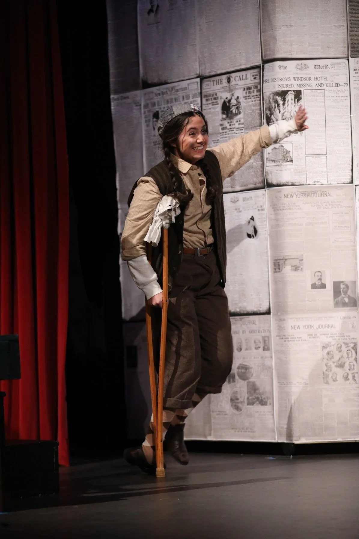 A young woman on stage dressed in vintage clothing, wearing a newsboy cap, with a backpack, holding a wooden cane, smiling and waving. The background features a wall with old newspaper pages and red stage curtains to the left.