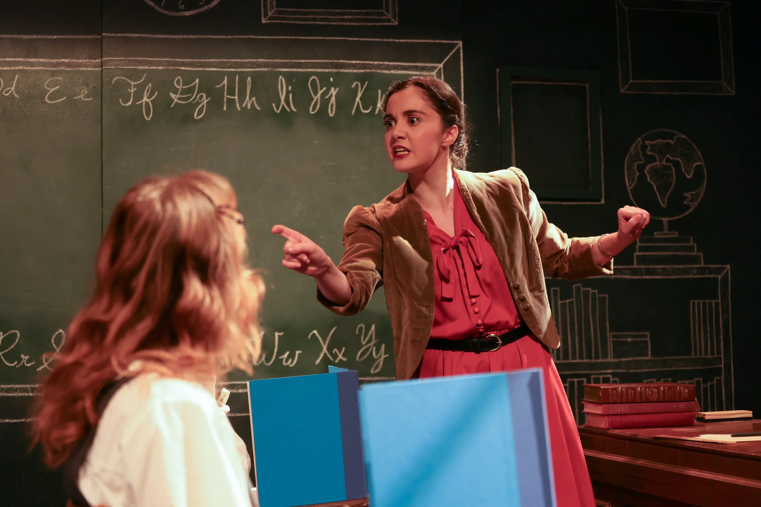 A teacher passionately instructs students in front of a chalkboard filled with mathematical and scientific formulas, with a stack of textbooks on a desk nearby.