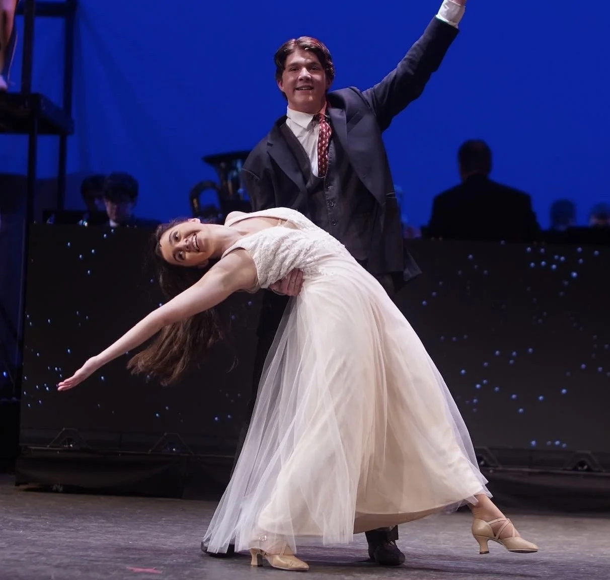 A couple dance on stage with the woman in a flowing pink gown and the man in a dark suit, performing a graceful pose with the woman leaning back and the man holding her, under stage lighting with a blue background.