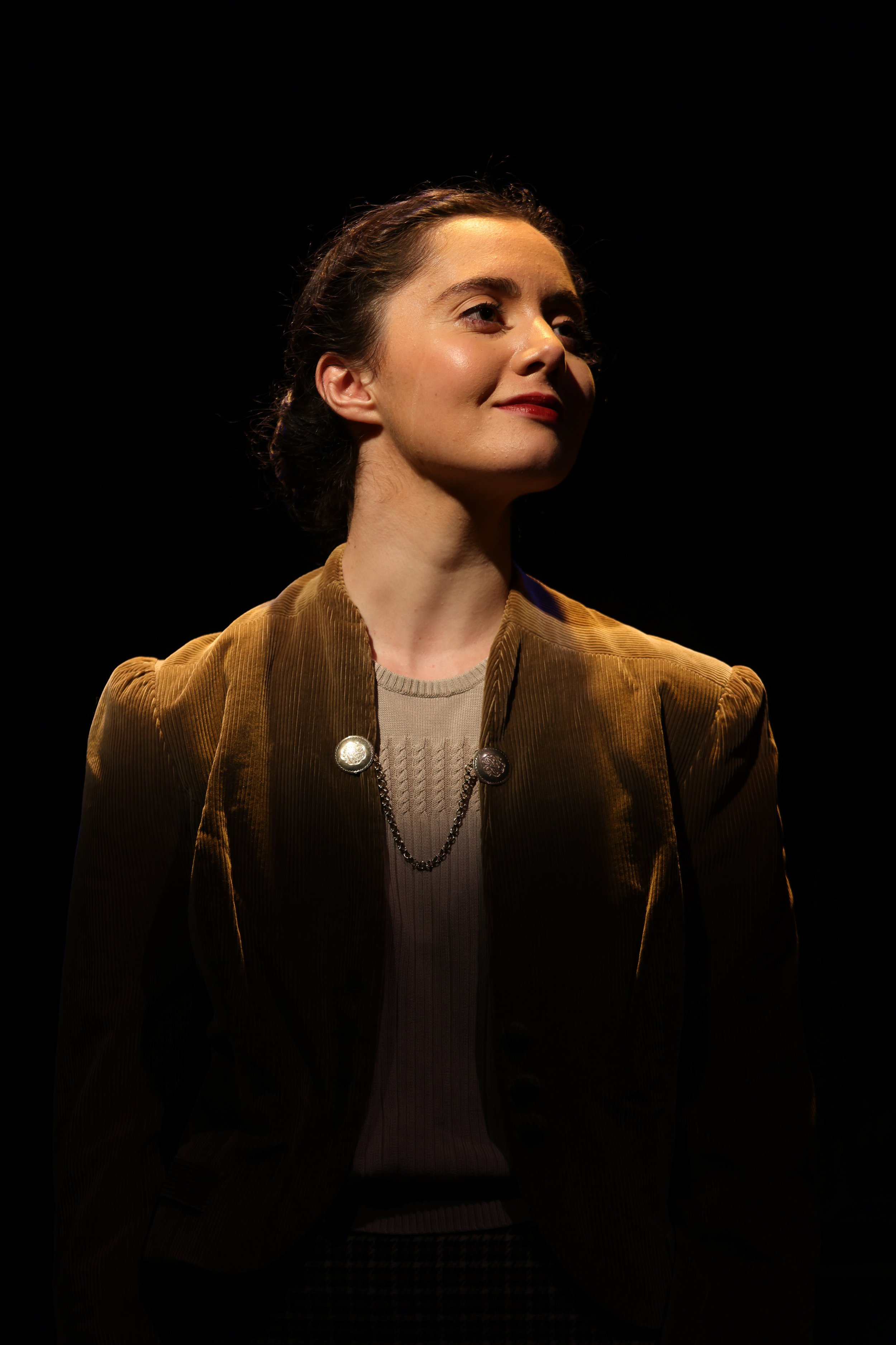 A woman with dark hair and fair skin is shown in profile, smiling slightly, in a dark setting with a spotlight on her face. She is wearing a brown velvet jacket, a gray top, and a silver chain necklace.
