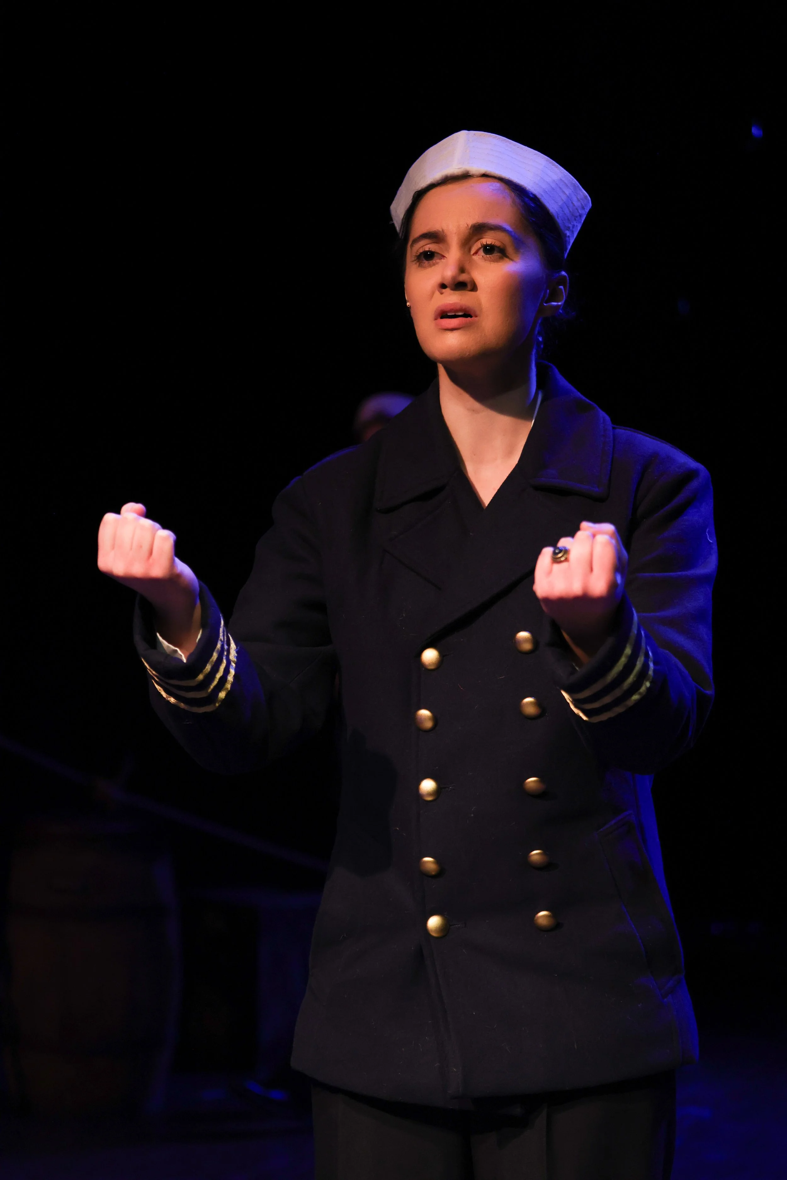 Woman dressed as a military officer with a white cap and dark coat, making a fist gesture on stage with a dark background.
