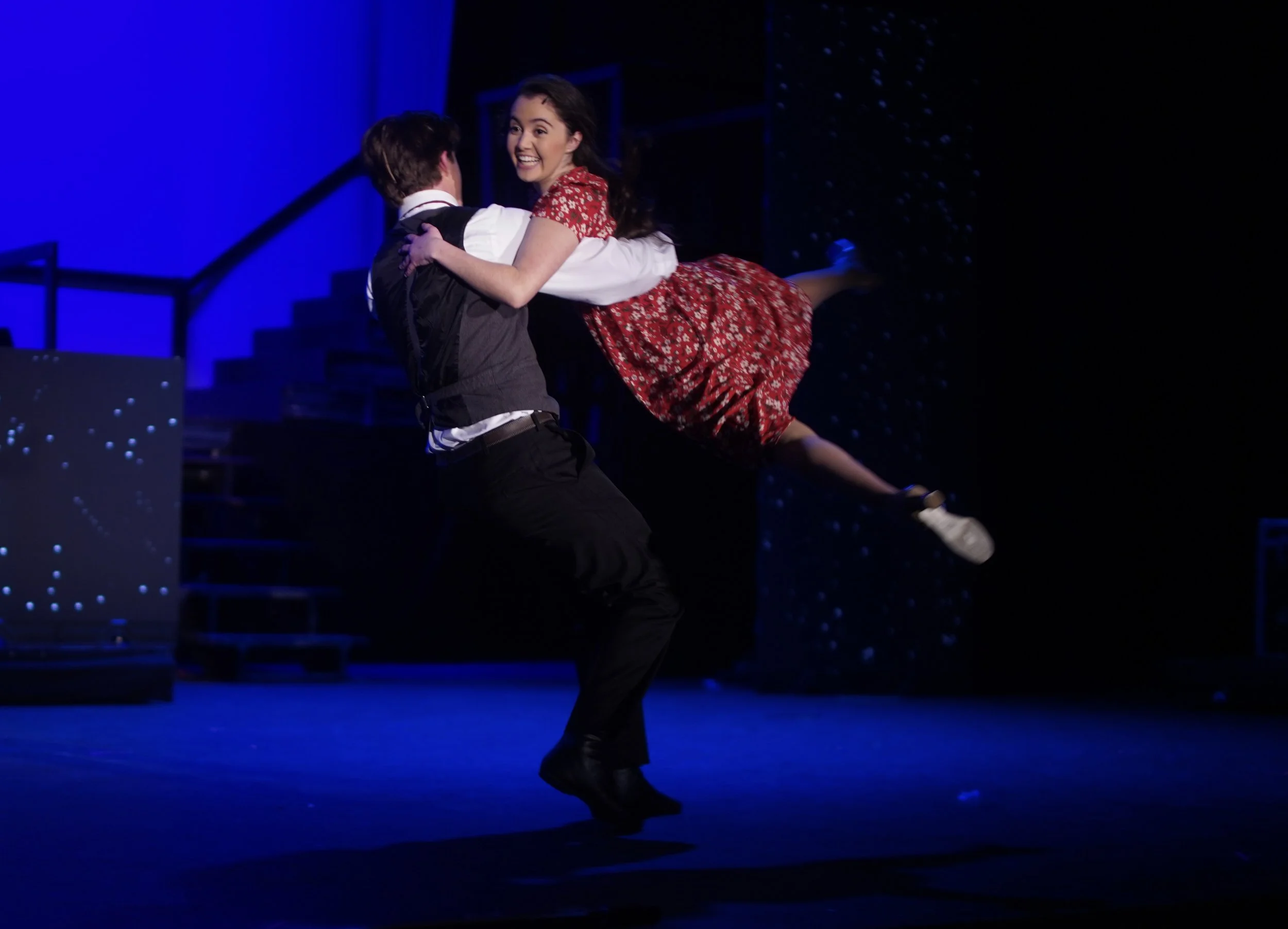 A young woman and a young man are dancing on a stage, with the woman lifted off the ground by the man. The woman is smiling brightly and wearing a red dress with a floral pattern. The stage is lit with blue lighting, and the background features stair