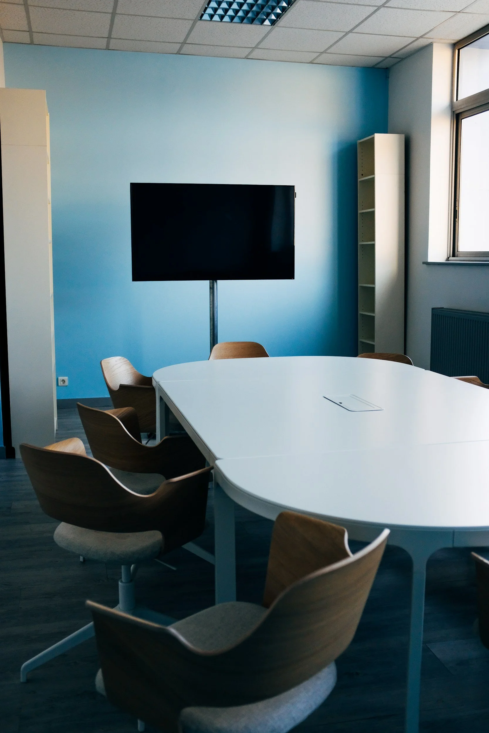 An office or conference room with a light blue wall, a black television on a stand, a window letting in natural light, and a white table surrounded by wooden chairs.