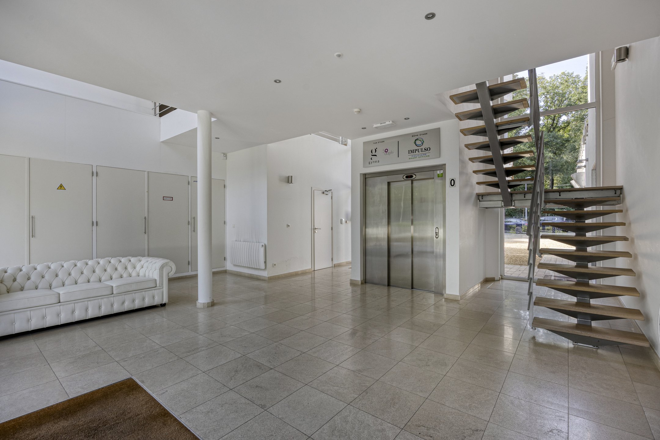 Interior of a modern building lobby with white walls, tiled floor, a staircase with wooden steps and metal railing, a white tufted sofa, and an elevator with metallic doors. There are windows with greenery visible outside.