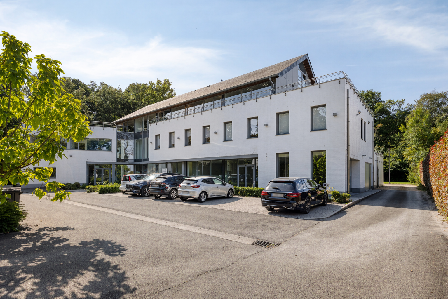 Modern white building with several parked cars in front, surrounded by greenery and trees under a blue sky.