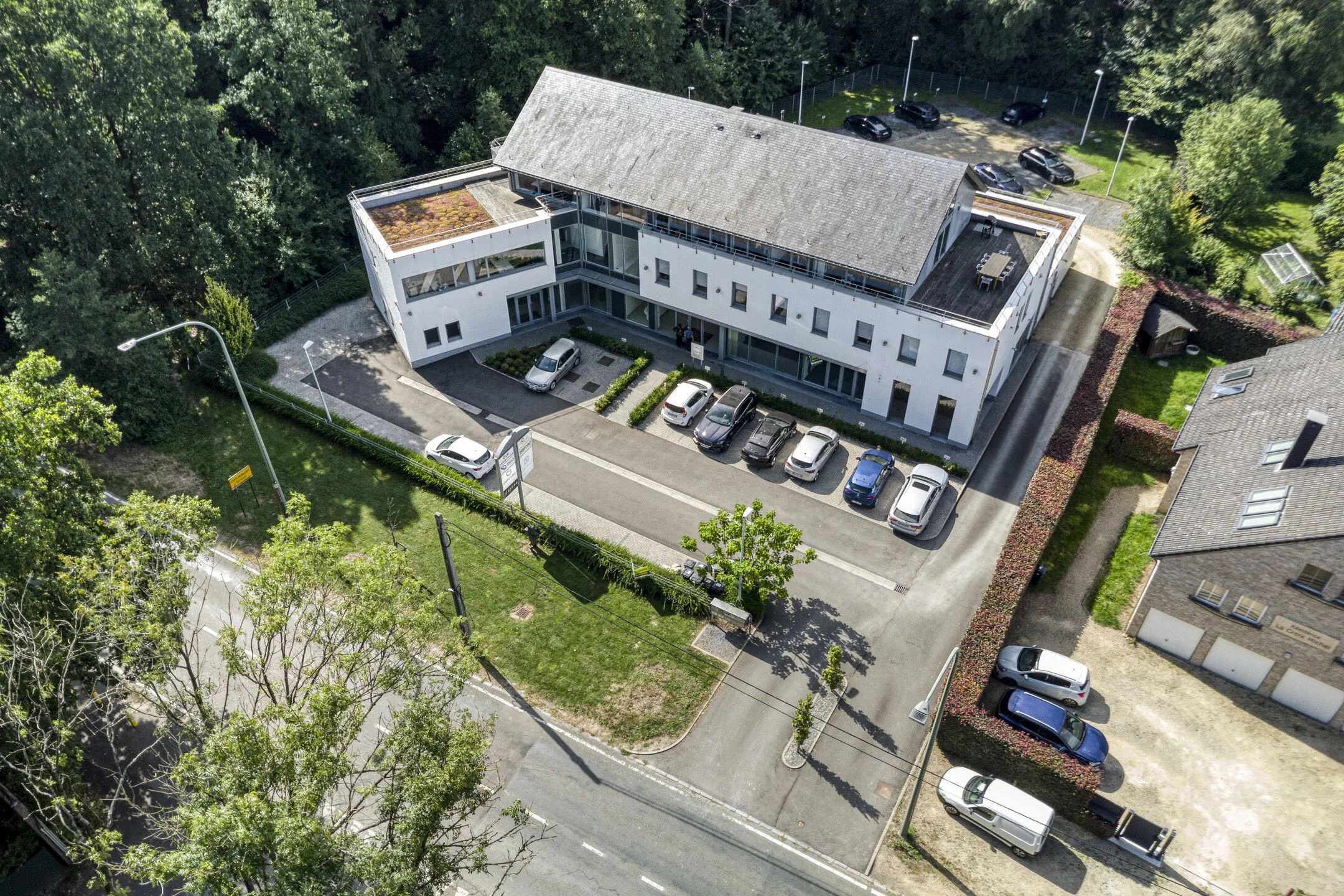An aerial view of a modern multi-story building surrounded by trees, with parking spaces and cars in front and behind, and a small garden with a greenhouse nearby.