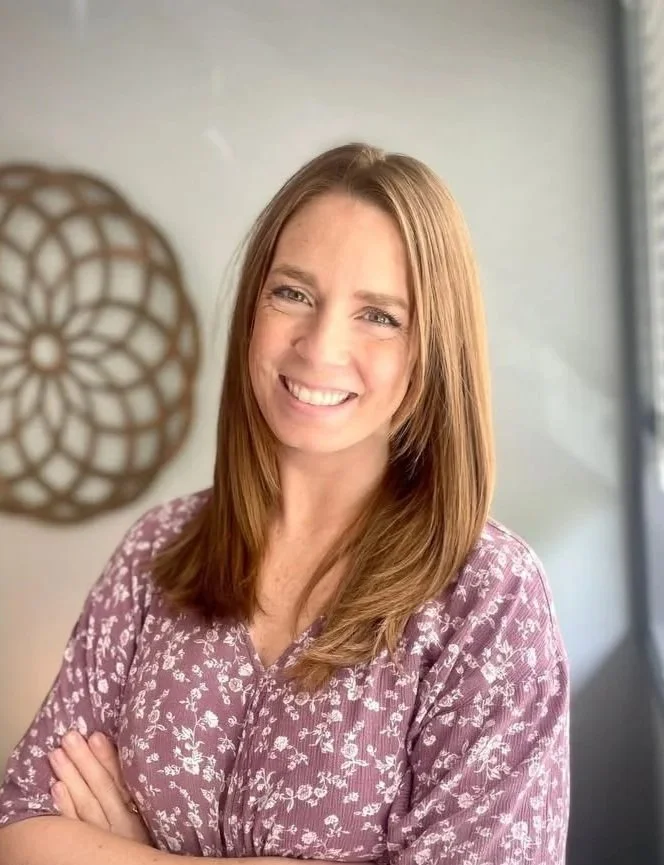Smiling woman standing in front of a light-colored wall with a decorative round wall art piece.