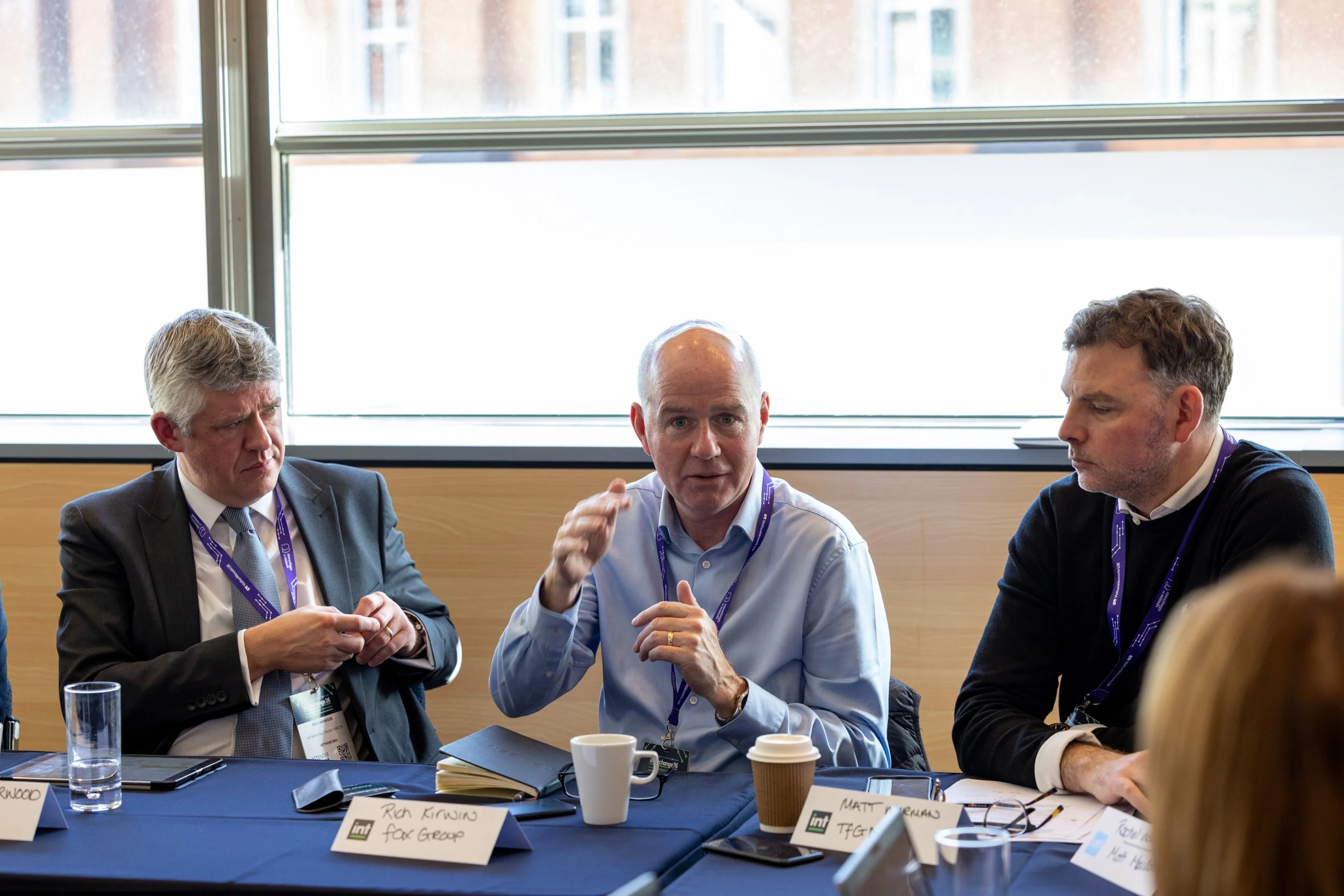 Three men sitting at a table engaged in conversation during a meeting or conference, with name tags and coffee cups in front of them.