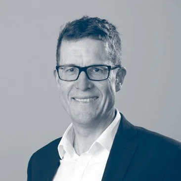 A man in a suit with glasses, smiling against a plain background.