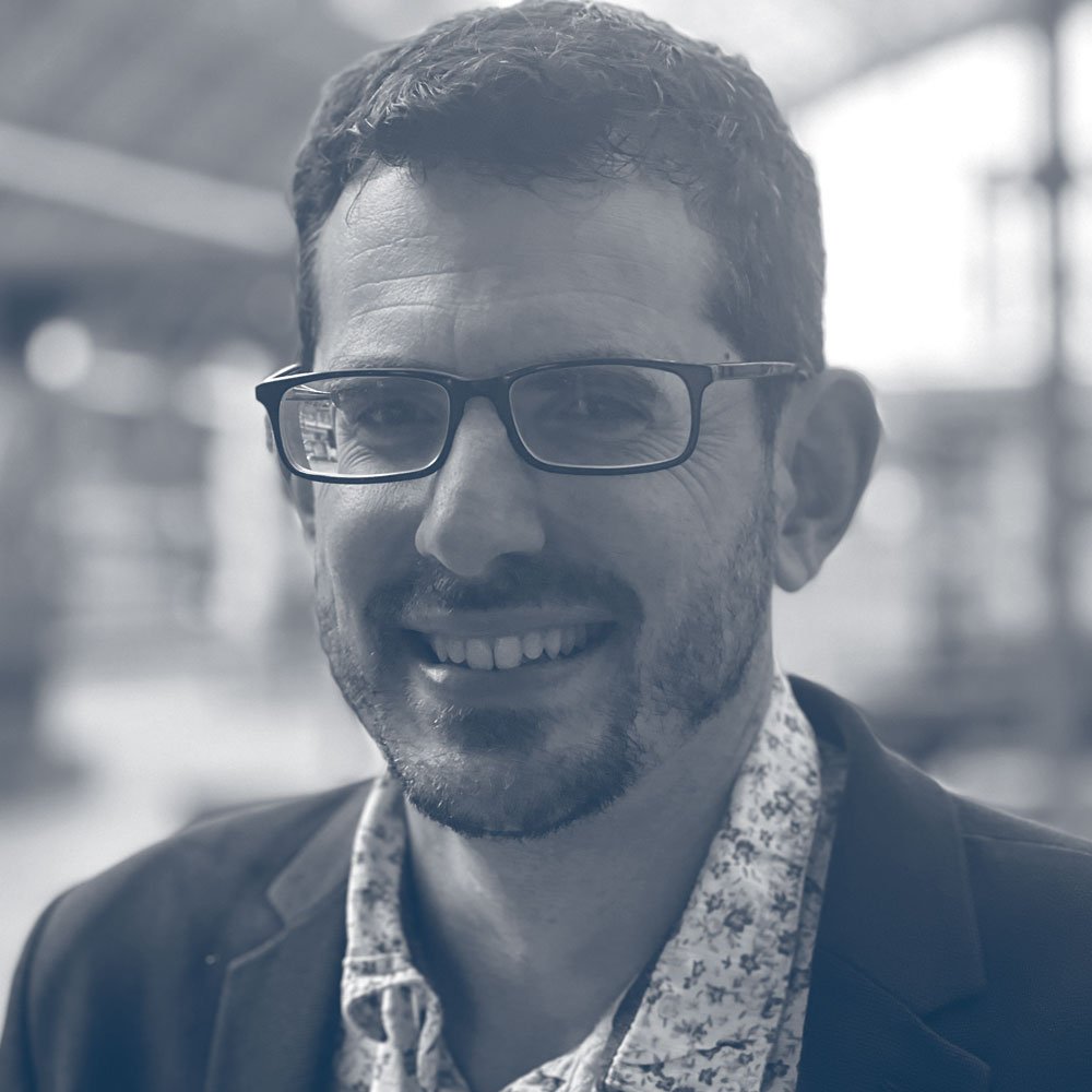 A man with glasses smiling indoors, wearing a blazer and a patterned shirt.