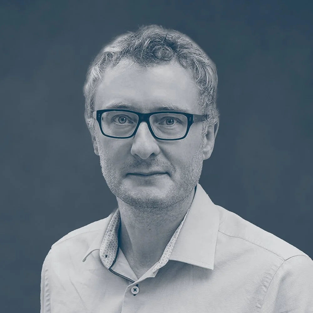 A middle-aged man with gray curly hair, wearing black glasses and a light-colored collared shirt, photo in grayscale.
