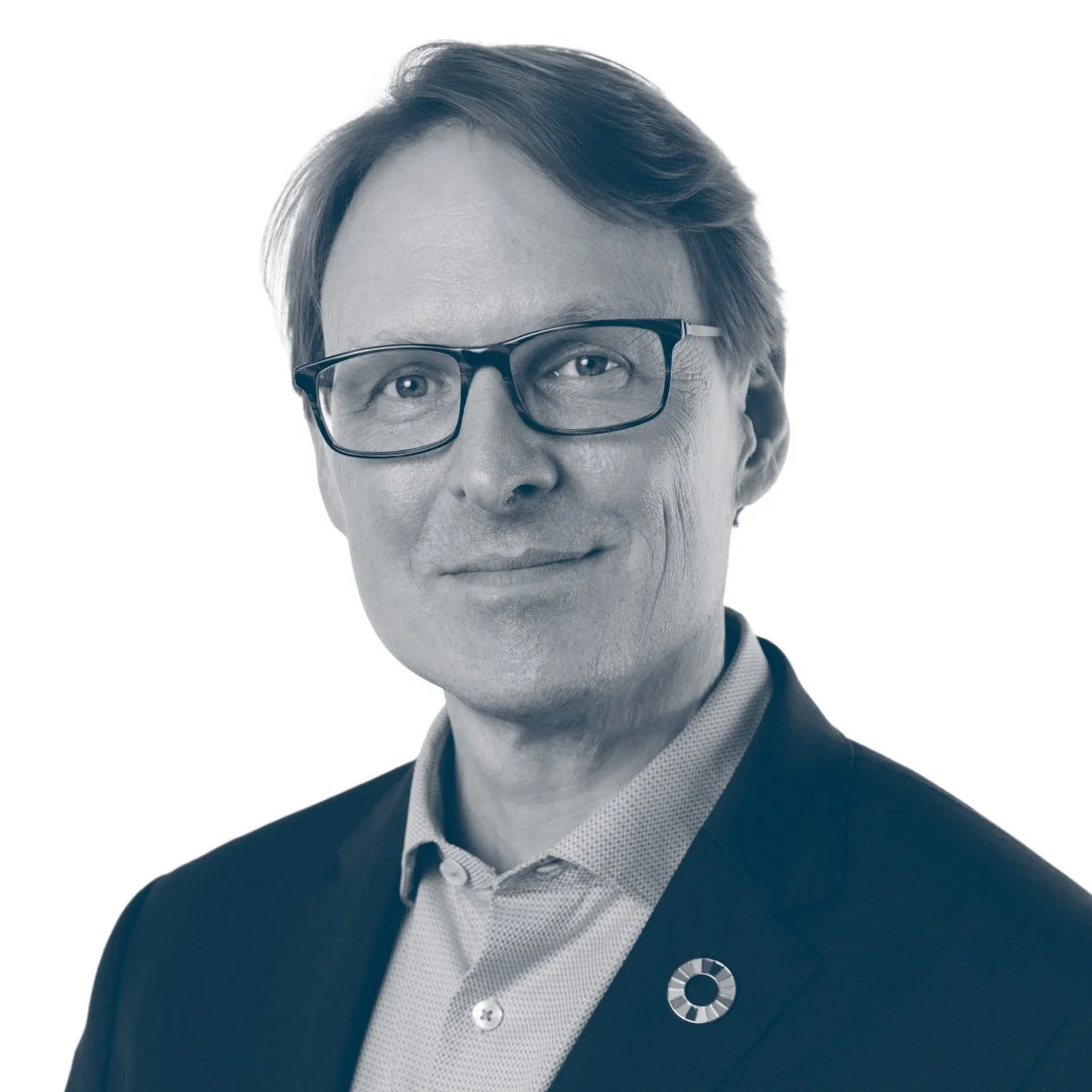 Black and white portrait of a middle-aged man wearing glasses, a suit jacket, and a collared shirt, smiling slightly, with a circular pin on his lapel.