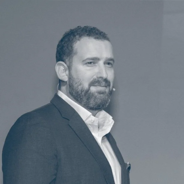 A man with a beard wearing a suit and white shirt, standing against a plain background.