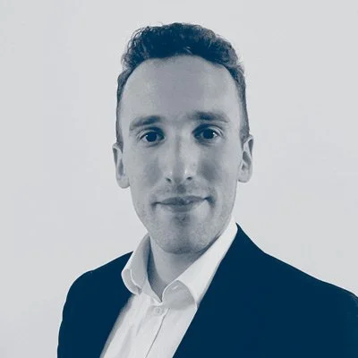 A grayscale portrait of a young man dressed in a white shirt and dark blazer, looking at the camera with a slight smile.