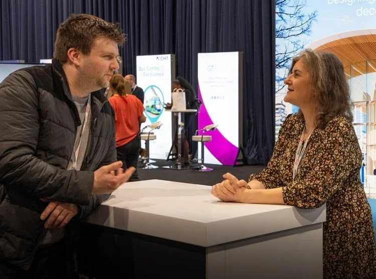 A man and woman engaged in conversation at a conference booth. The man is wearing a black jacket, and the woman is in a floral dress. Behind them are banners and other attendees.