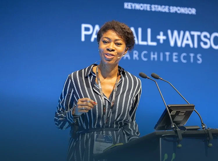 A woman speaking at a podium during a conference with a blue background. The backdrop displays text referencing a keynote speaker sponsored by Parametric + Watson Architects.