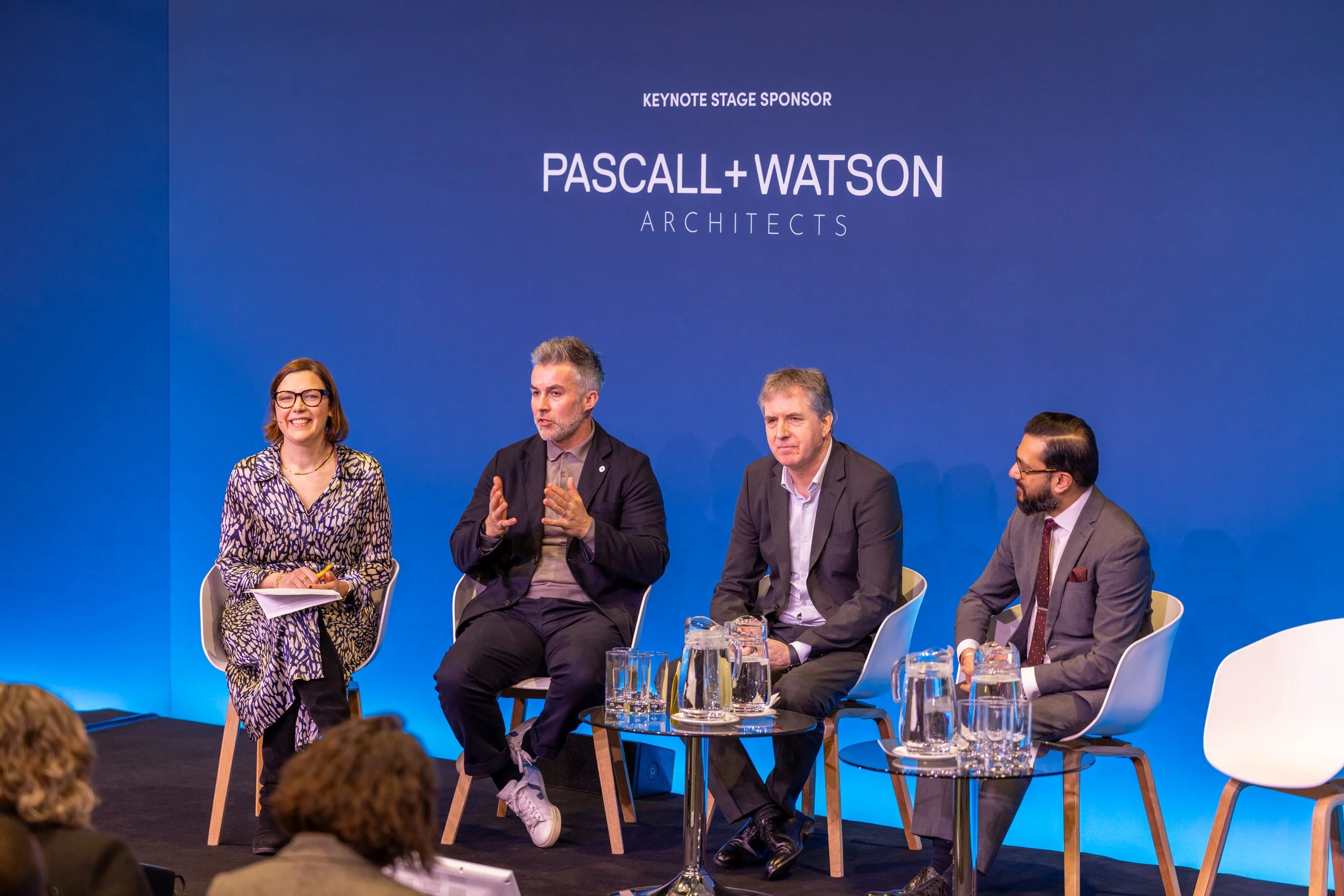 Panel discussion at an event with four individuals sitting and one standing woman, against a blue background with text 'Keynote Stage Sponsor Pascall + Watson Architects'.