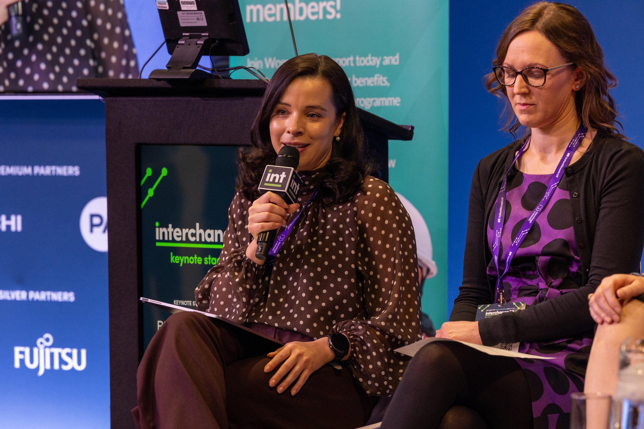 Two women sitting on a stage at a conference, one holding a microphone, with a blue and teal background with text and logos, and a monitor displaying the words "interchange keynote speaker".