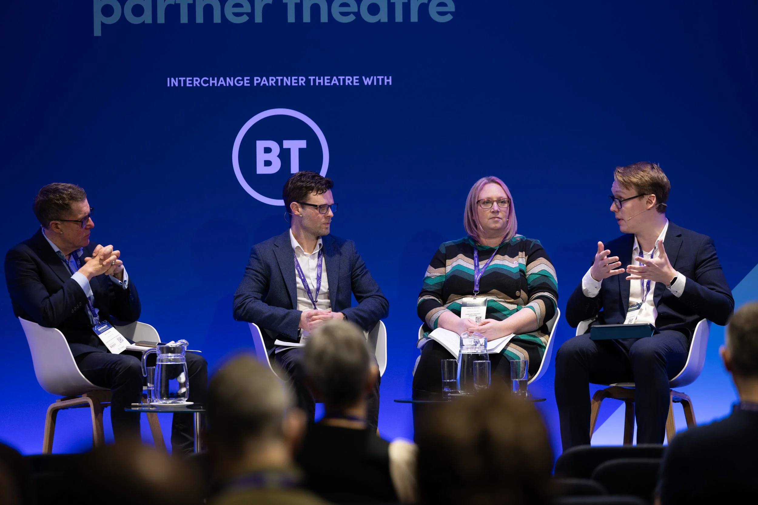 Four people seated on stage engaged in a panel discussion. There are glasses of water in front of them, and the background has a blue screen with text related to the event partner and logo.