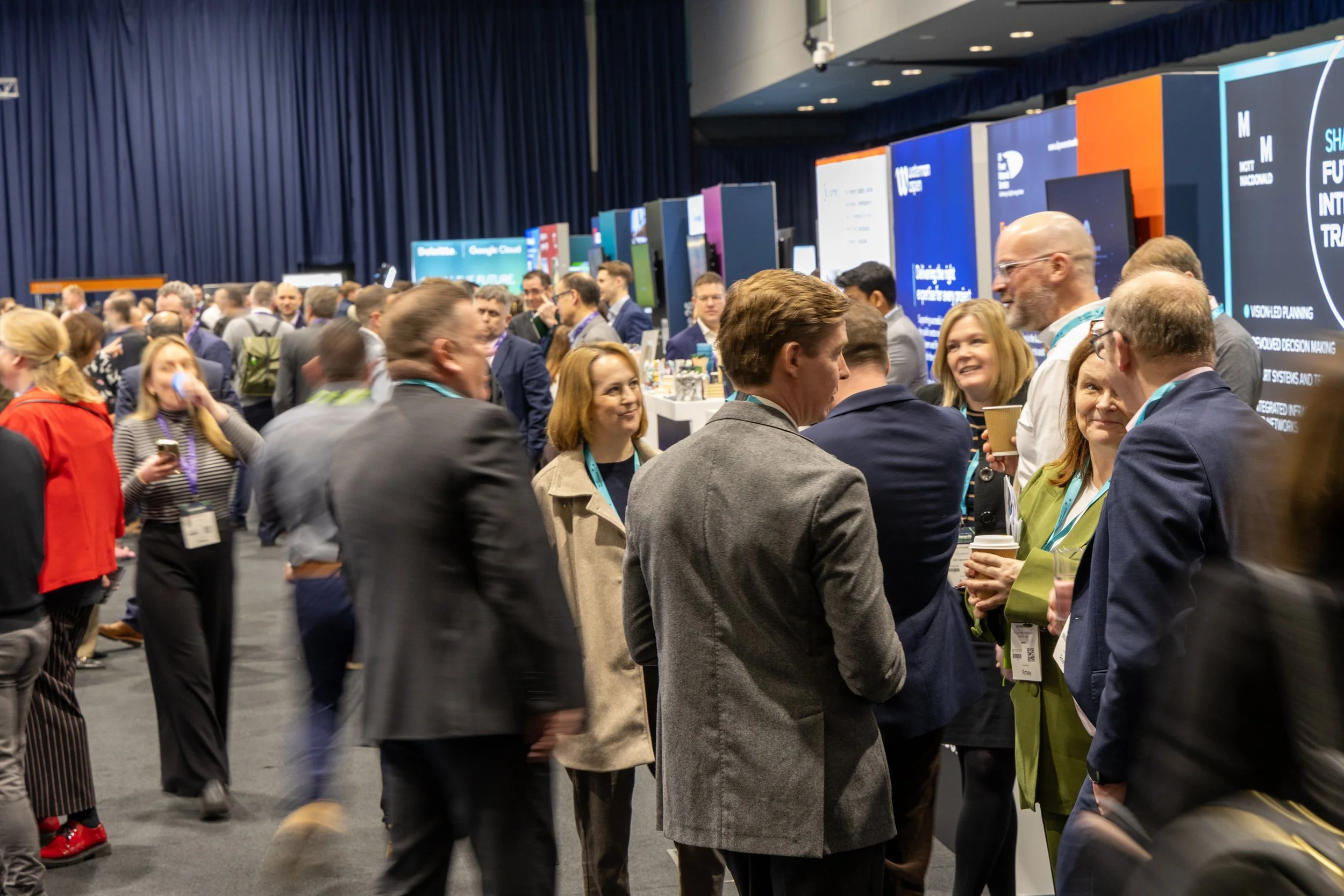 People attending a conference or expo, engaging in conversations, in a large hall with display booths and banners.