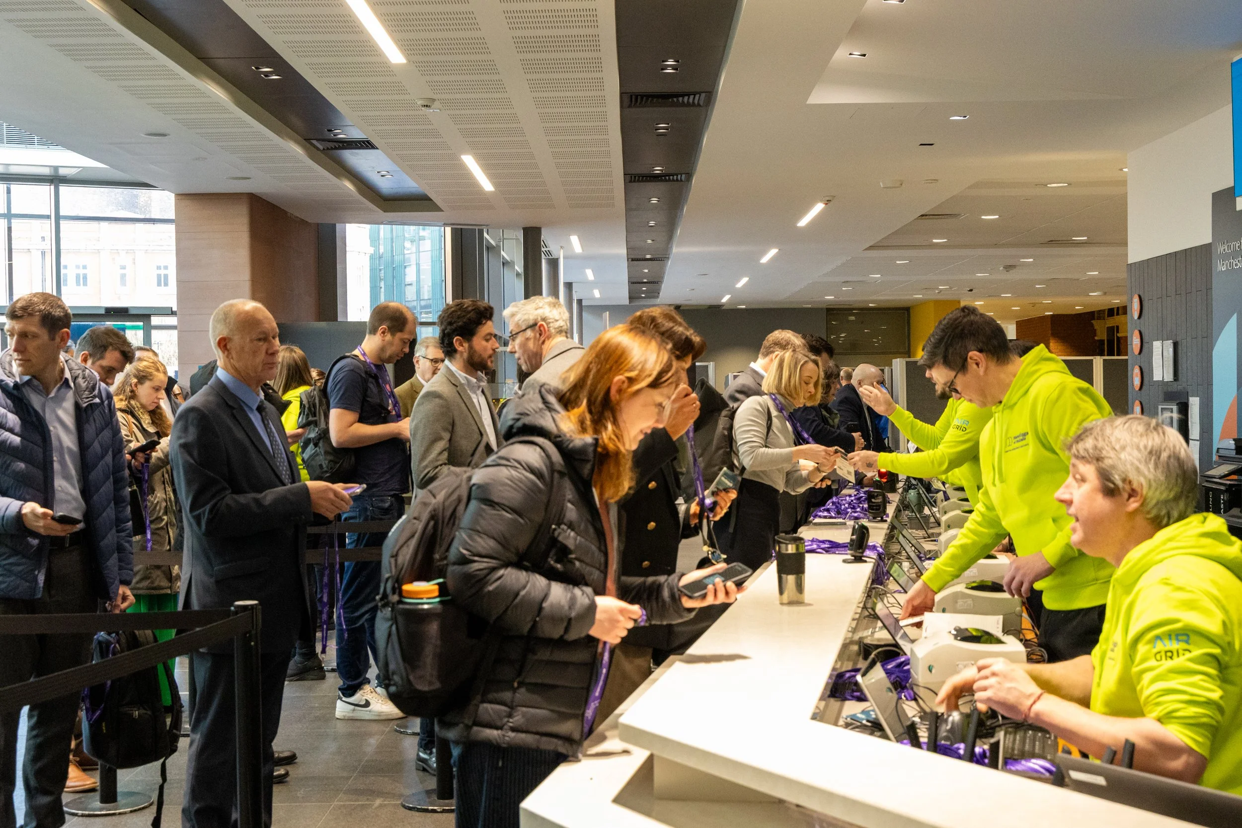 People standing in line at a registration or check-in counter at an indoor event, with staff in yellow shirts assisting and handing out materials.