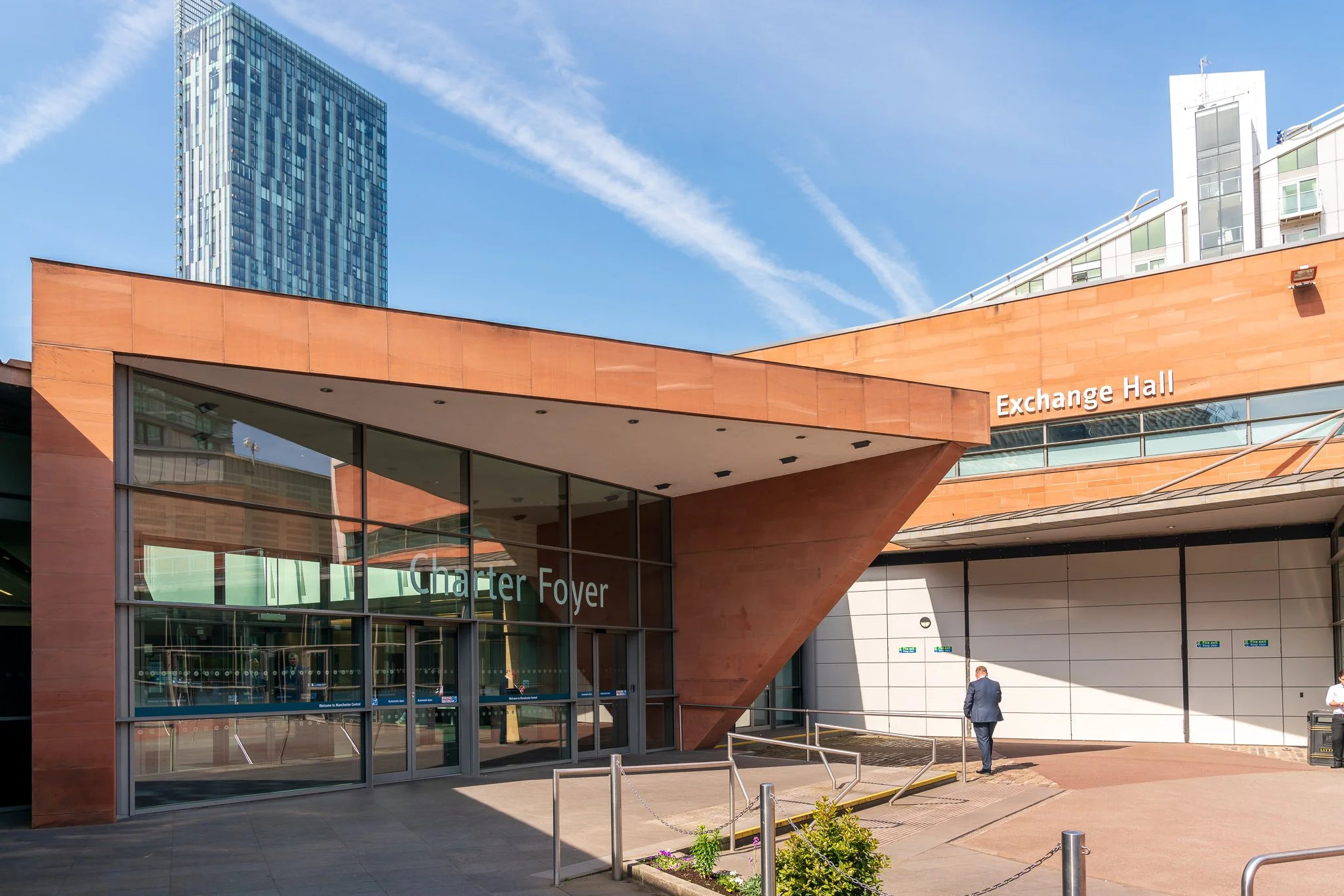 Entrance to a building labeled 'Charter Foyer' and 'Exchange Hall,' with a glass facade, a man in a suit walking away, and a city skyline with tall buildings in the background.