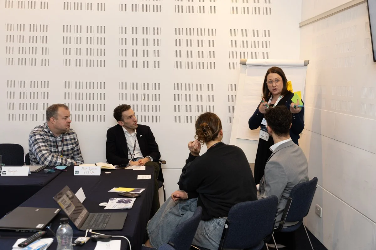 A woman standing next to a flip chart, speaking to a group of four people seated around a black conference table in a meeting room.
