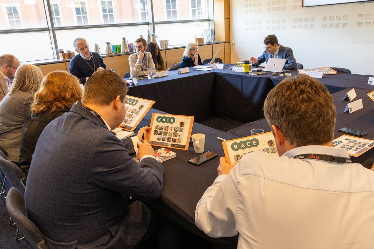 Group of business people sitting around a conference table, reading Race event information, in a well-lit room with large windows.