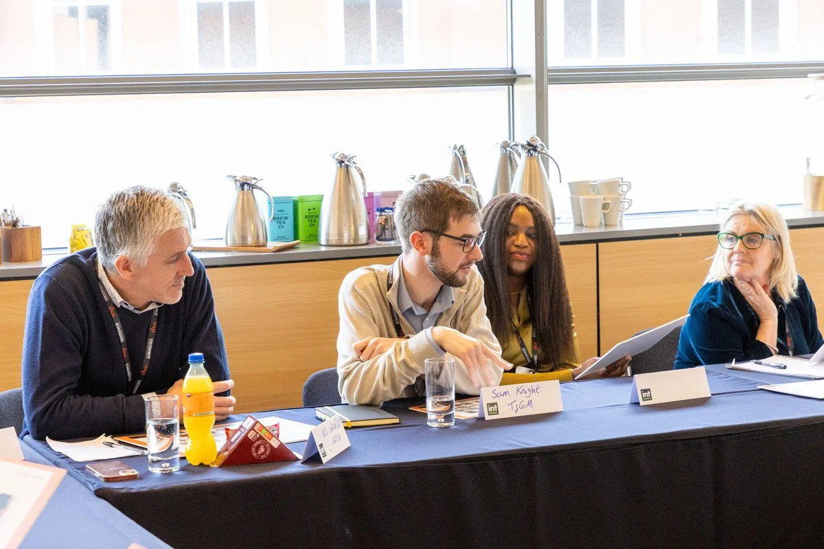 Four people sitting at a conference table in a room with large windows, engaged in discussion, with name tags, notebooks, and beverages on the table.